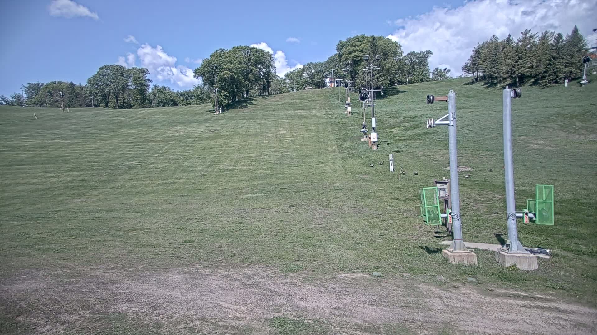 A grassy ski slope with a chairlift and trees in the background under a partly cloudy sky.