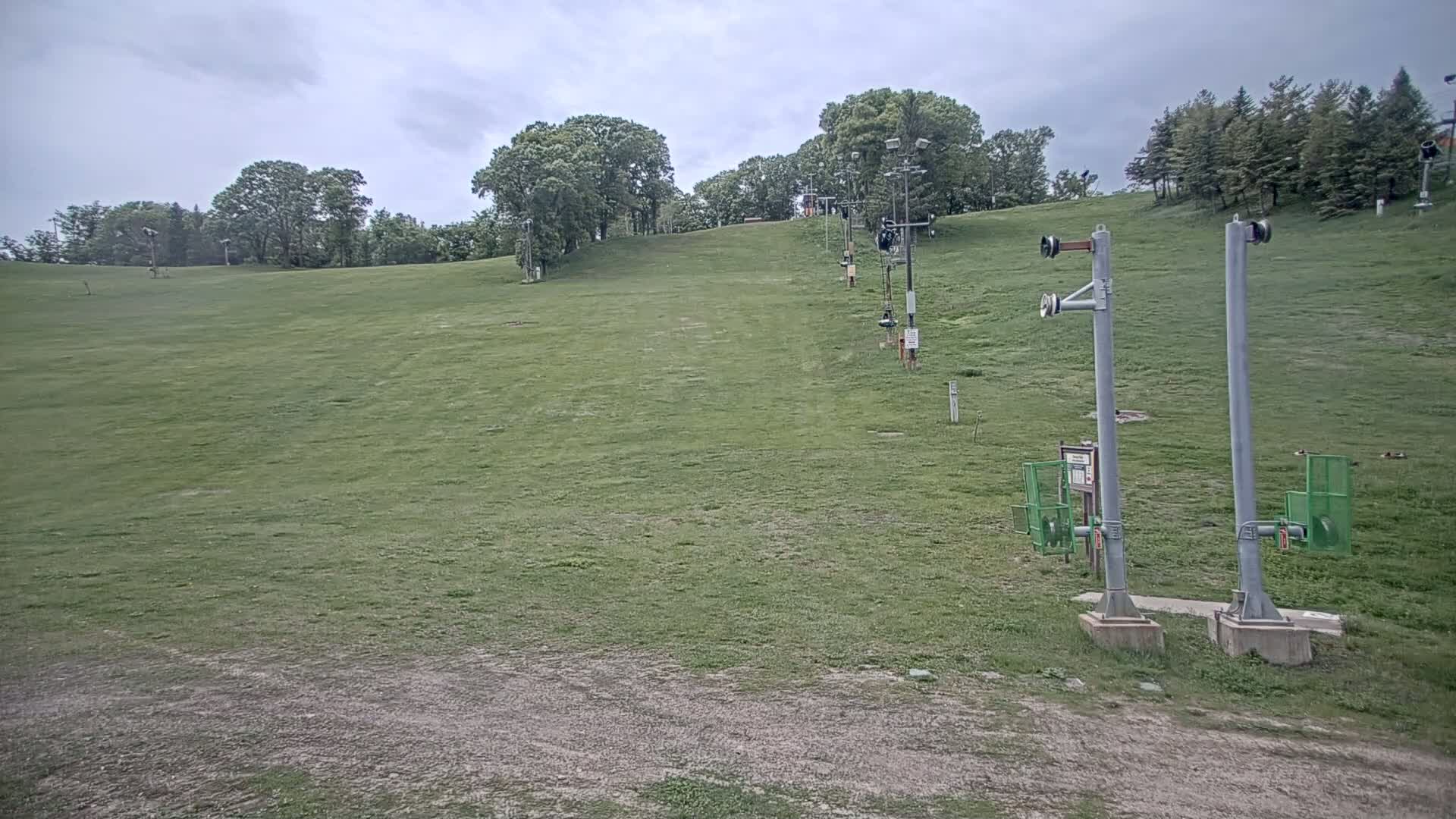 A grassy ski slope with a chairlift in the background under a cloudy sky.