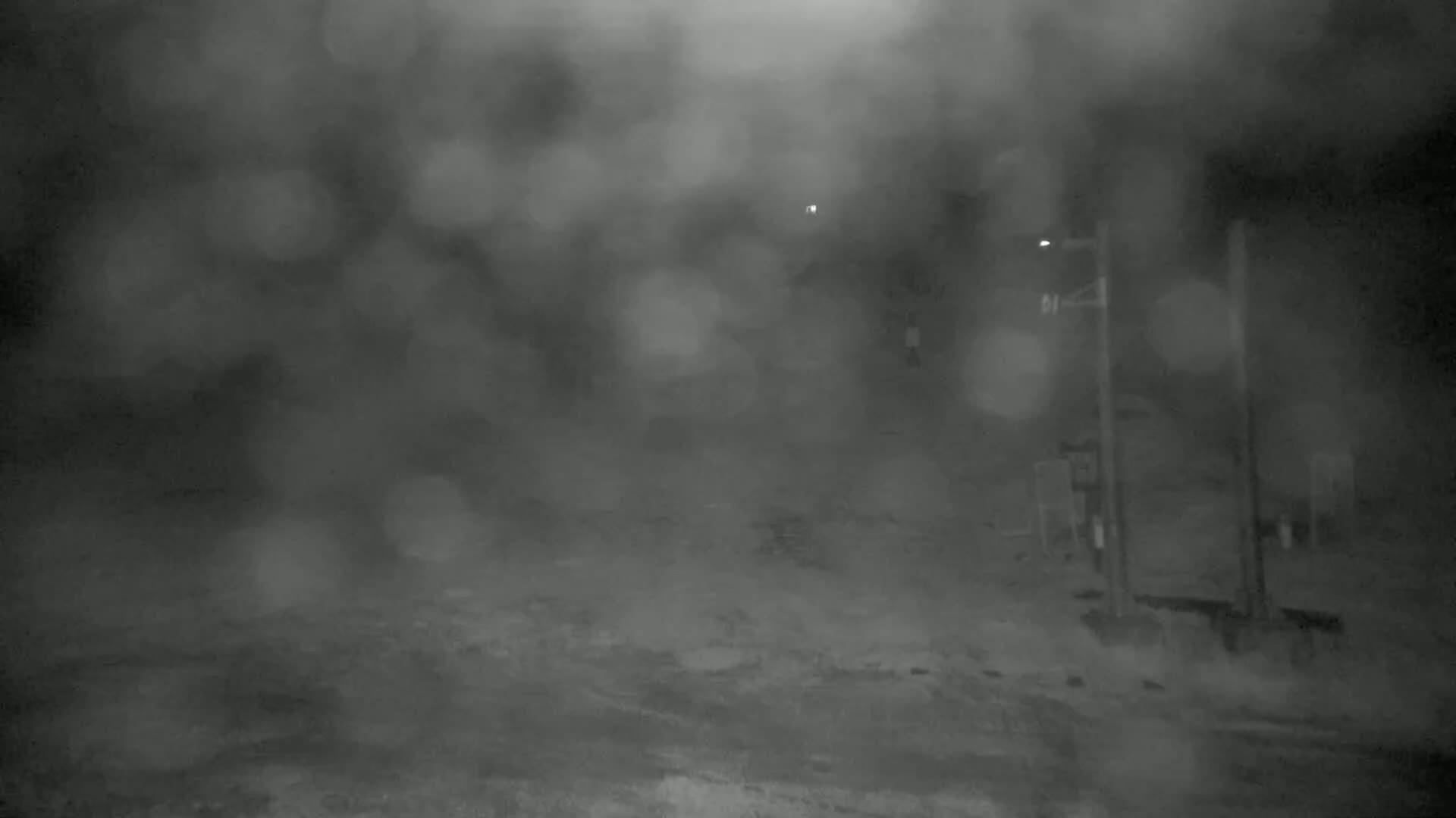 A blurry nighttime scene shows a mostly snow-covered area with a utility pole and faint lights in the distance, under heavy snow or fog.