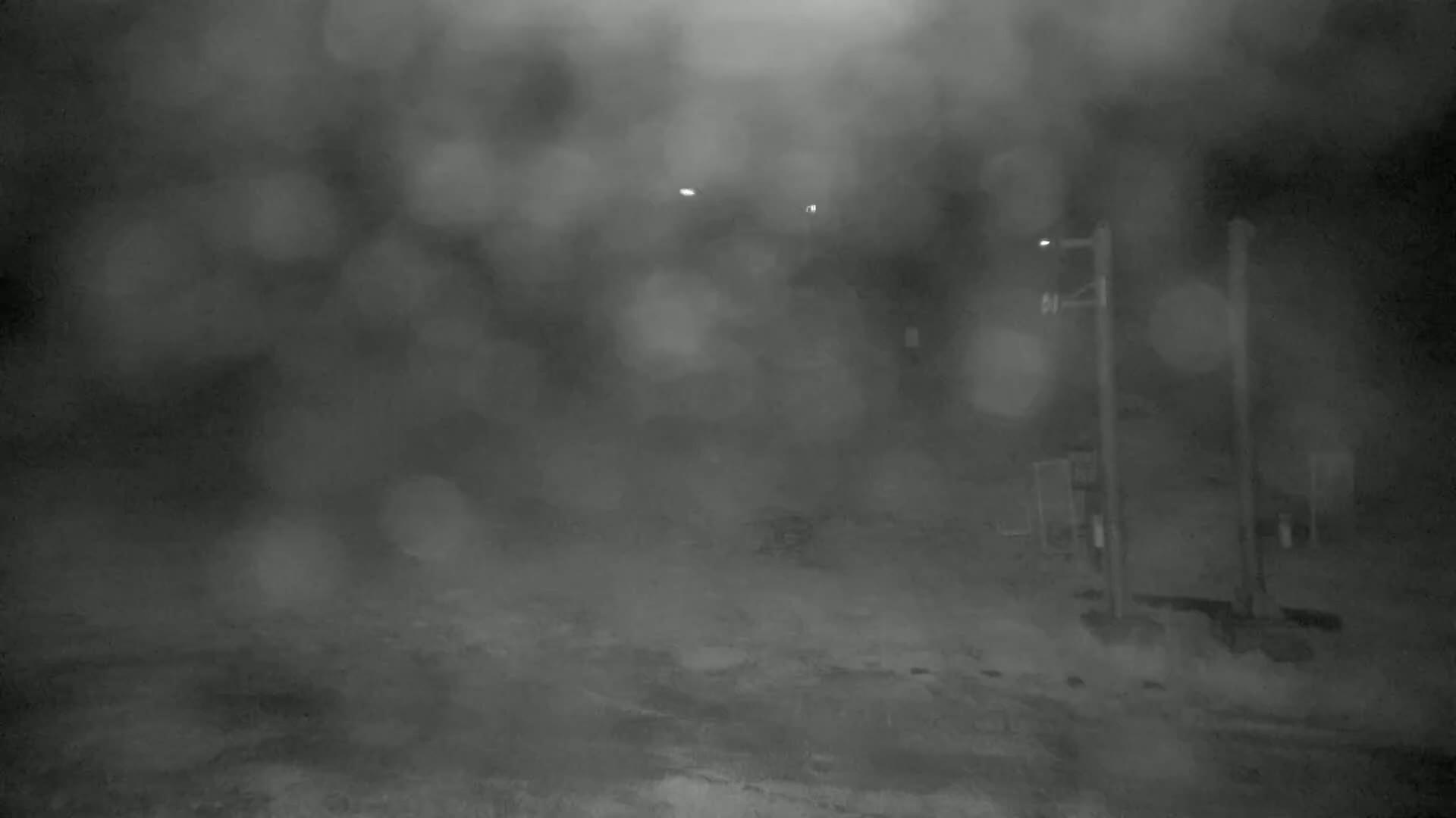 A blurry nighttime scene of an outdoor area shrouded in heavy snow or fog, featuring indistinct structures and a partially visible utility pole.