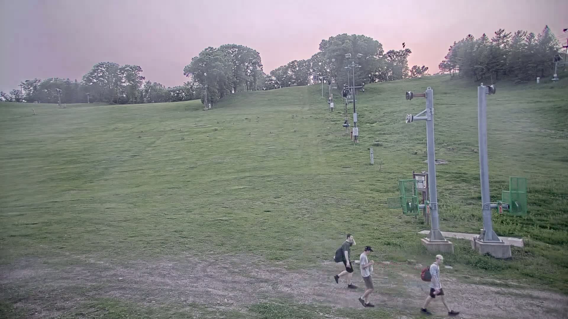 Three people walk across a grassy hillside under a pale pink sky near a ski lift.