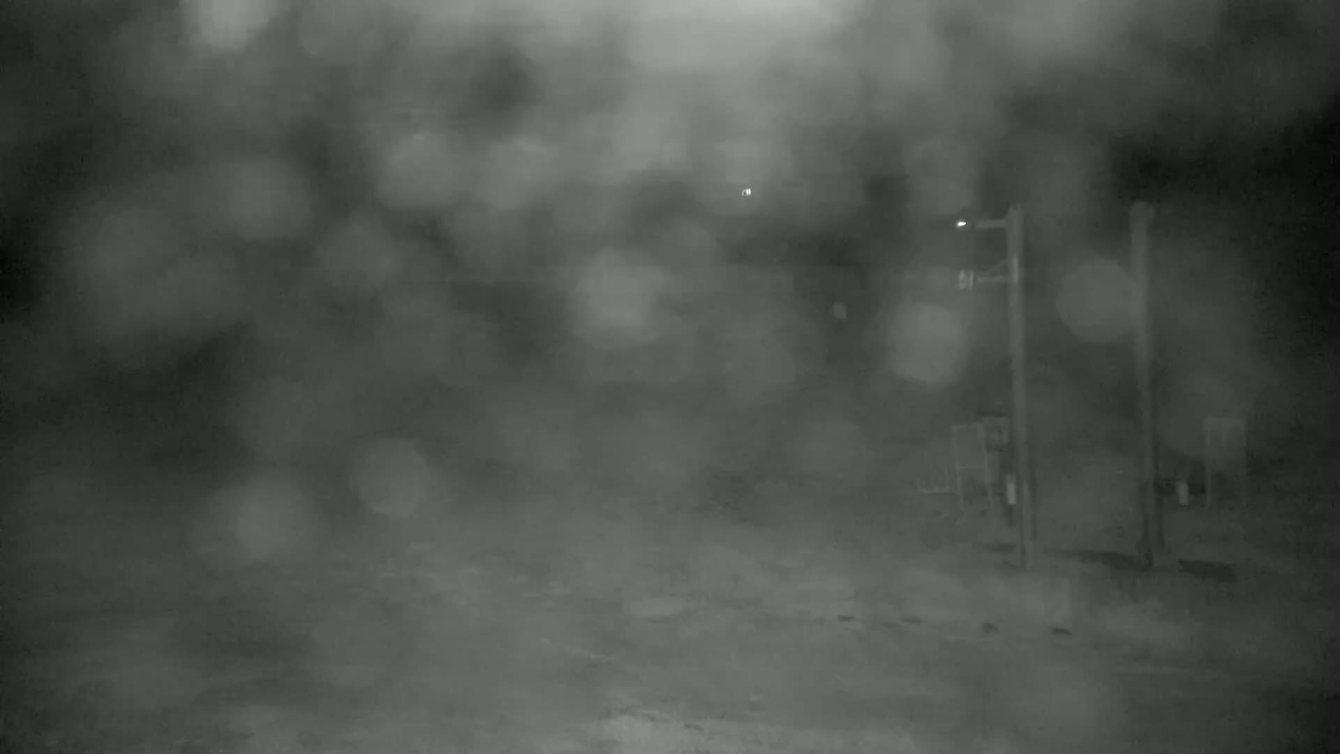 A blurry, nighttime outdoor scene shows a road with indistinct objects and poles, obscured by heavy rain or fog.