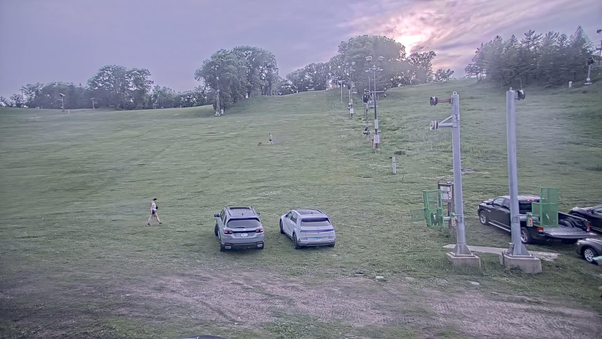 A grassy hill with a ski lift, two parked SUVs, a person walking a dog, and another person walking, all under a partly cloudy sunset sky.