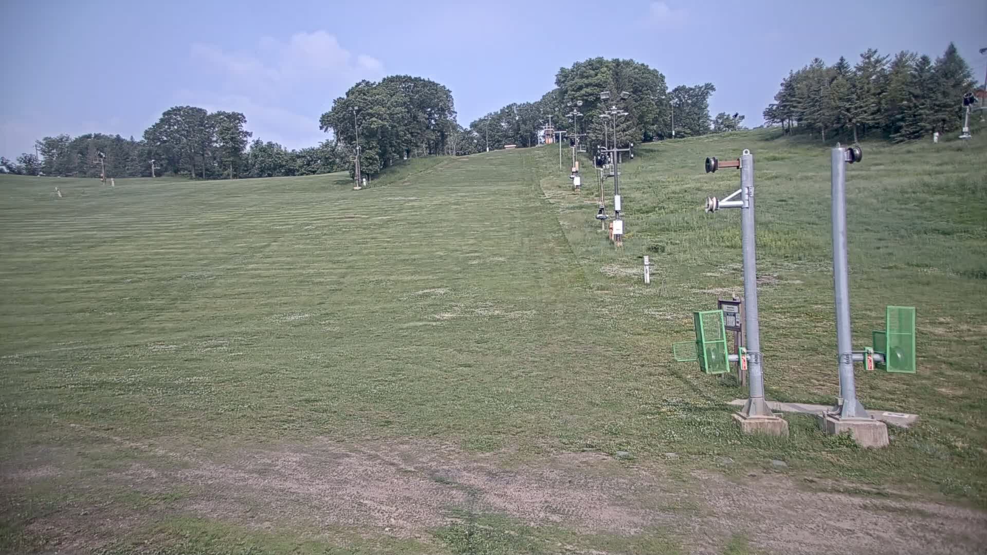 A grassy hill with a ski lift extends upward towards a line of trees under a mostly clear sky.