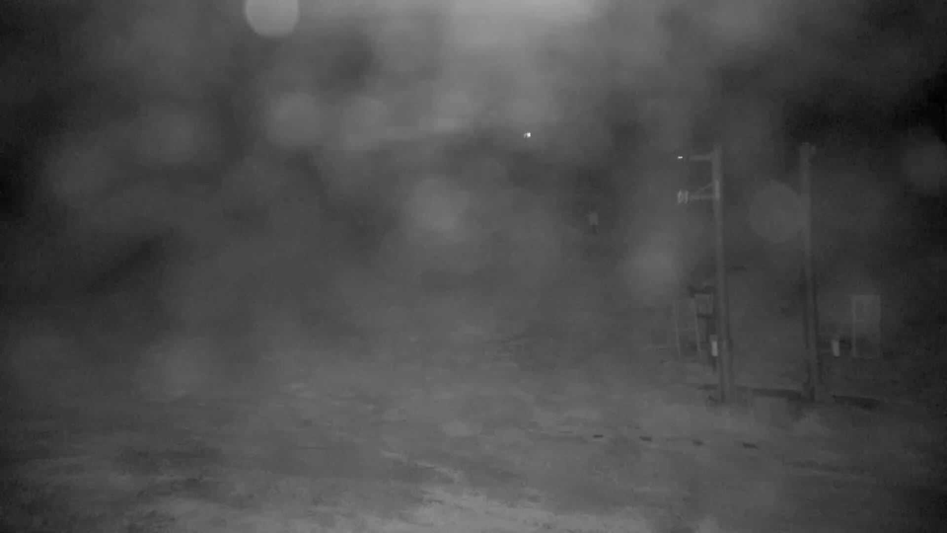 A blurry, nighttime outdoor scene shows a mostly empty, snow-covered area with some indistinct structures and poles under heavy snowfall.