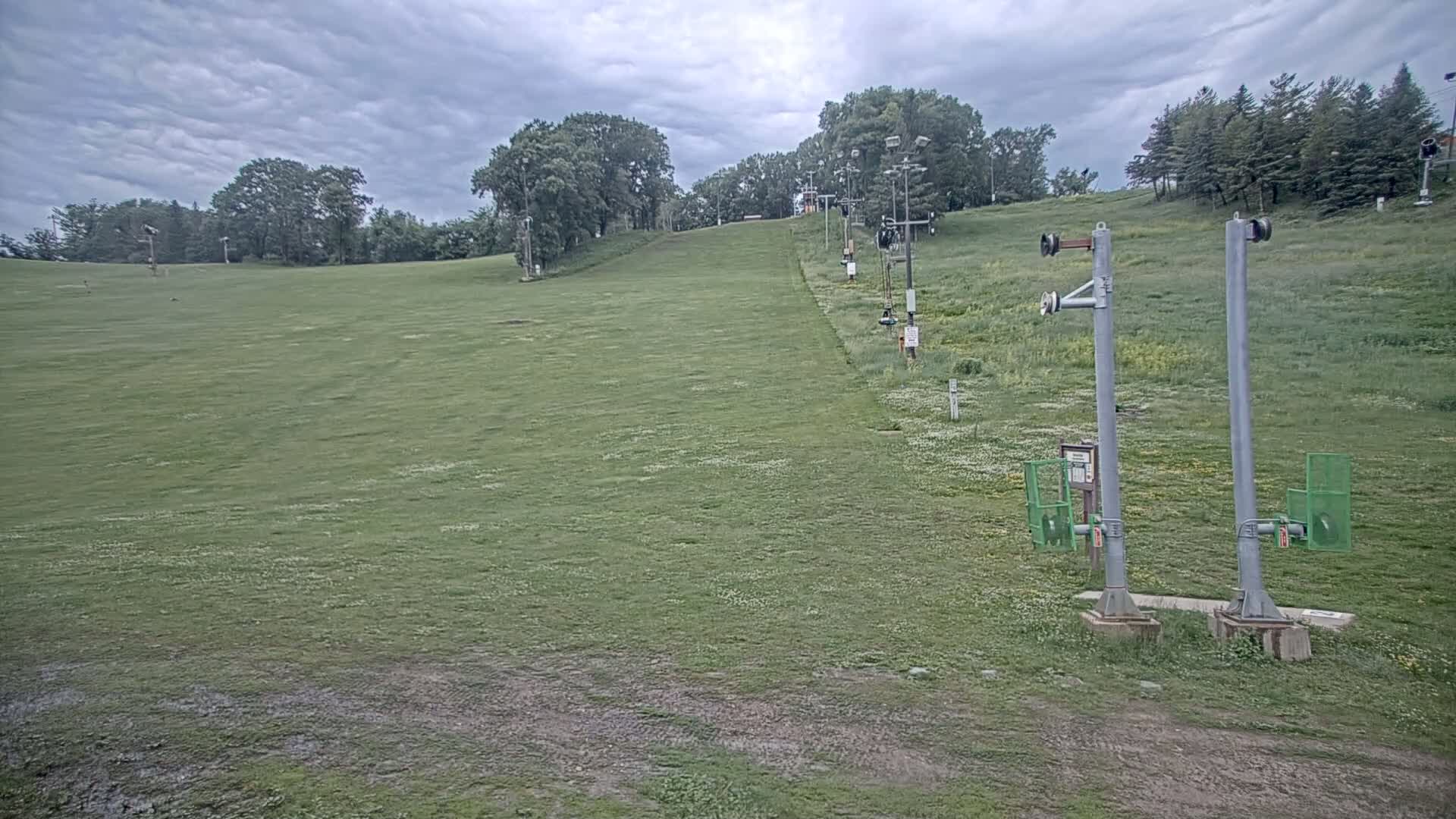 A grassy ski slope with a chairlift in the background under a cloudy sky.