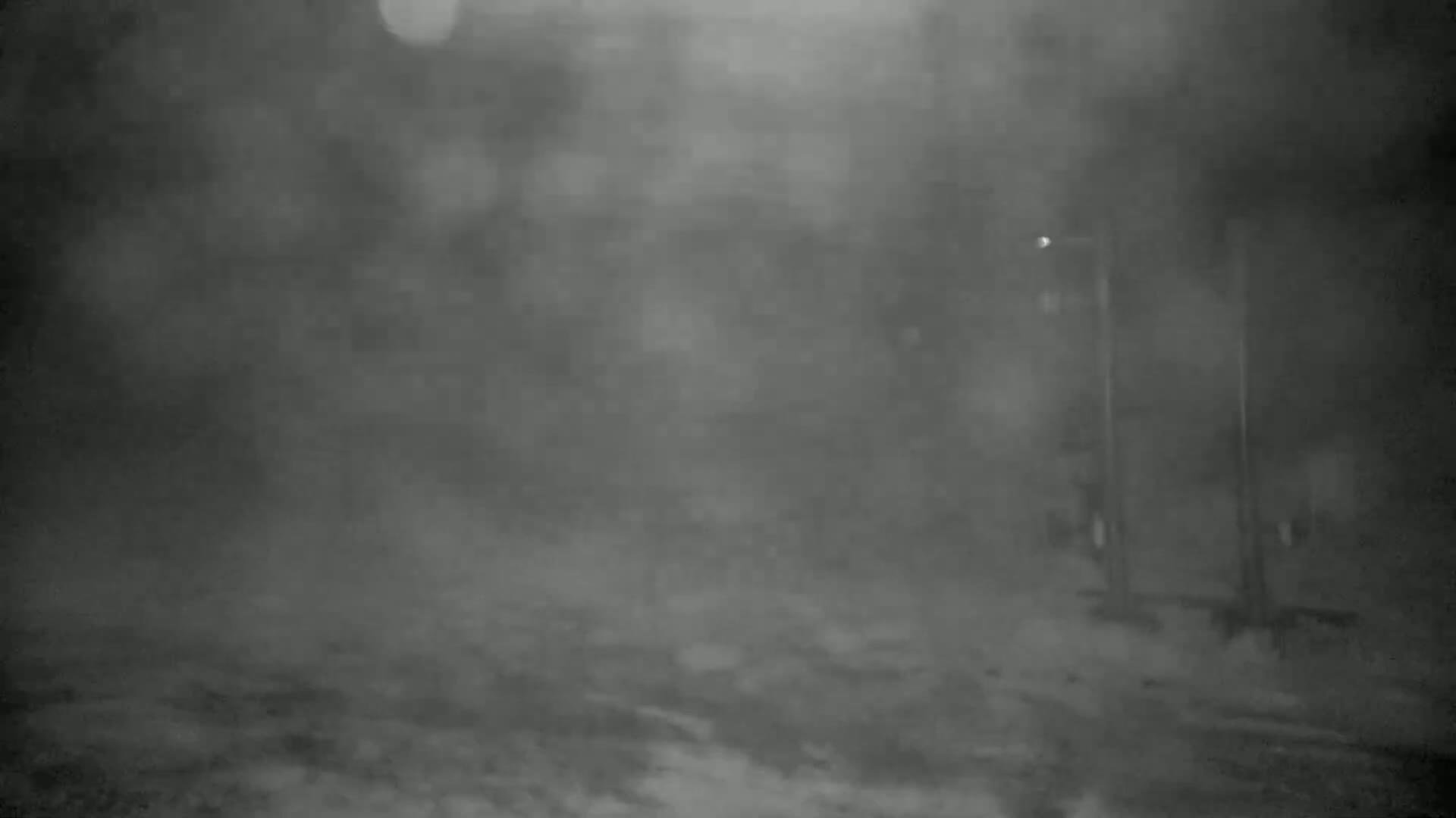 A blurry, nighttime image shows a snow-covered ground with indistinct structures and poles in the background under heavy snowfall.