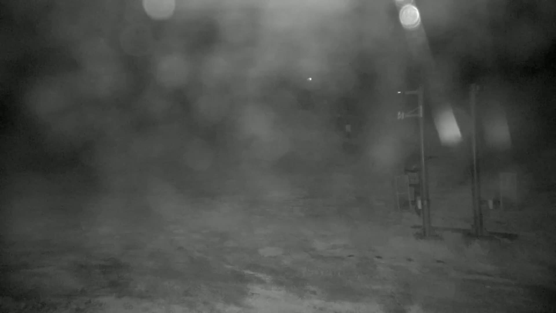 A blurry, nighttime image shows a snow-covered, possibly empty road with indistinct structures and posts in the background, under heavy snowfall.