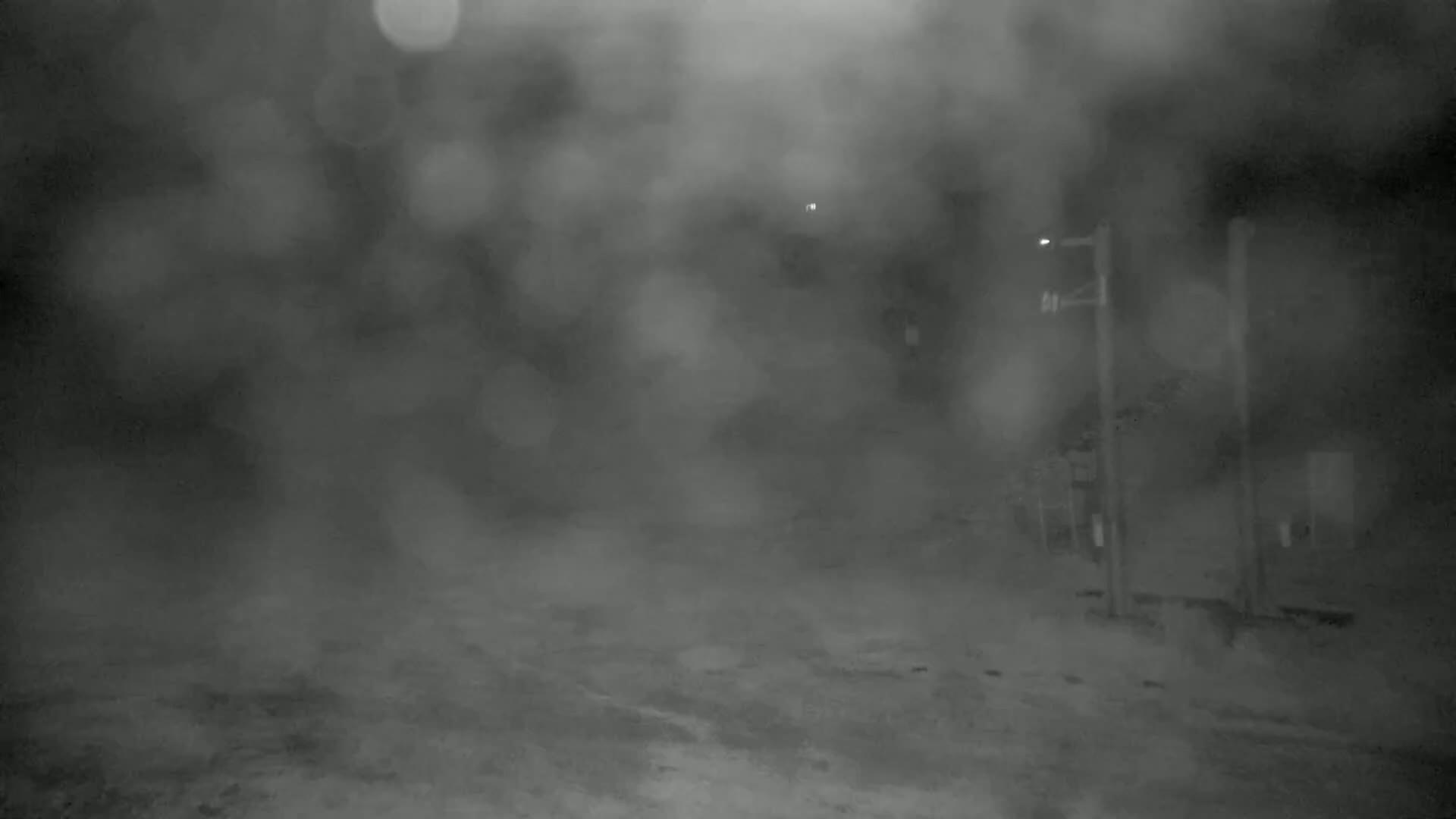 A blurry, nighttime outdoor scene shows a road and indistinct structures shrouded in heavy fog or blowing snow.