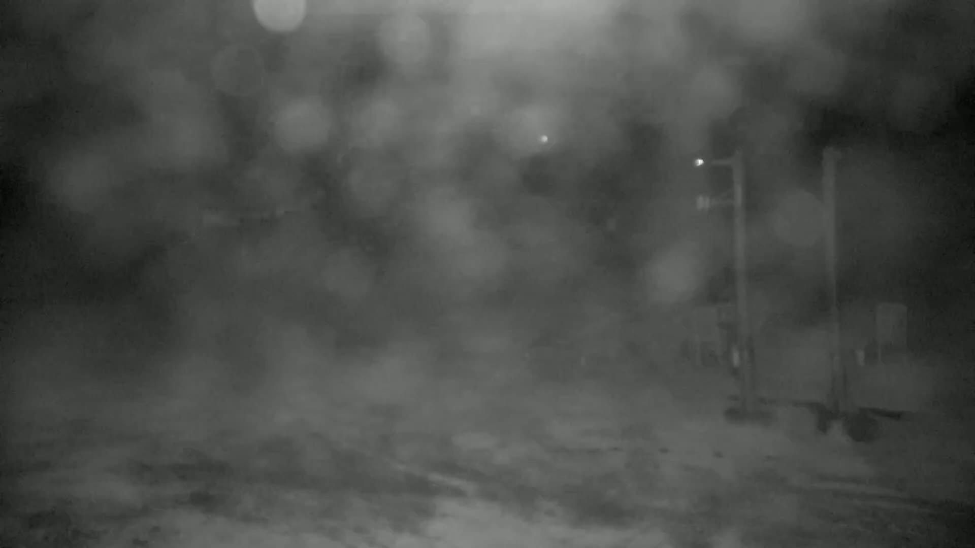 A blurry, nighttime image shows a dimly lit, snow-covered area with indistinct structures and a light post, obscured by heavy snowfall.