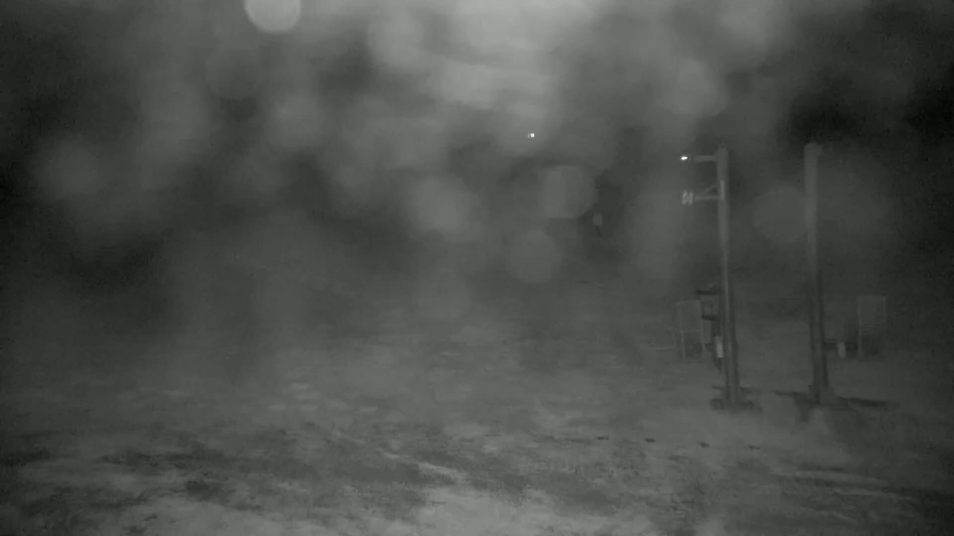 A blurry nighttime scene of a sparsely lit, possibly snowy or dusty, area with a tall post and some indistinct structures in the background, under heavy precipitation or a dense fog.