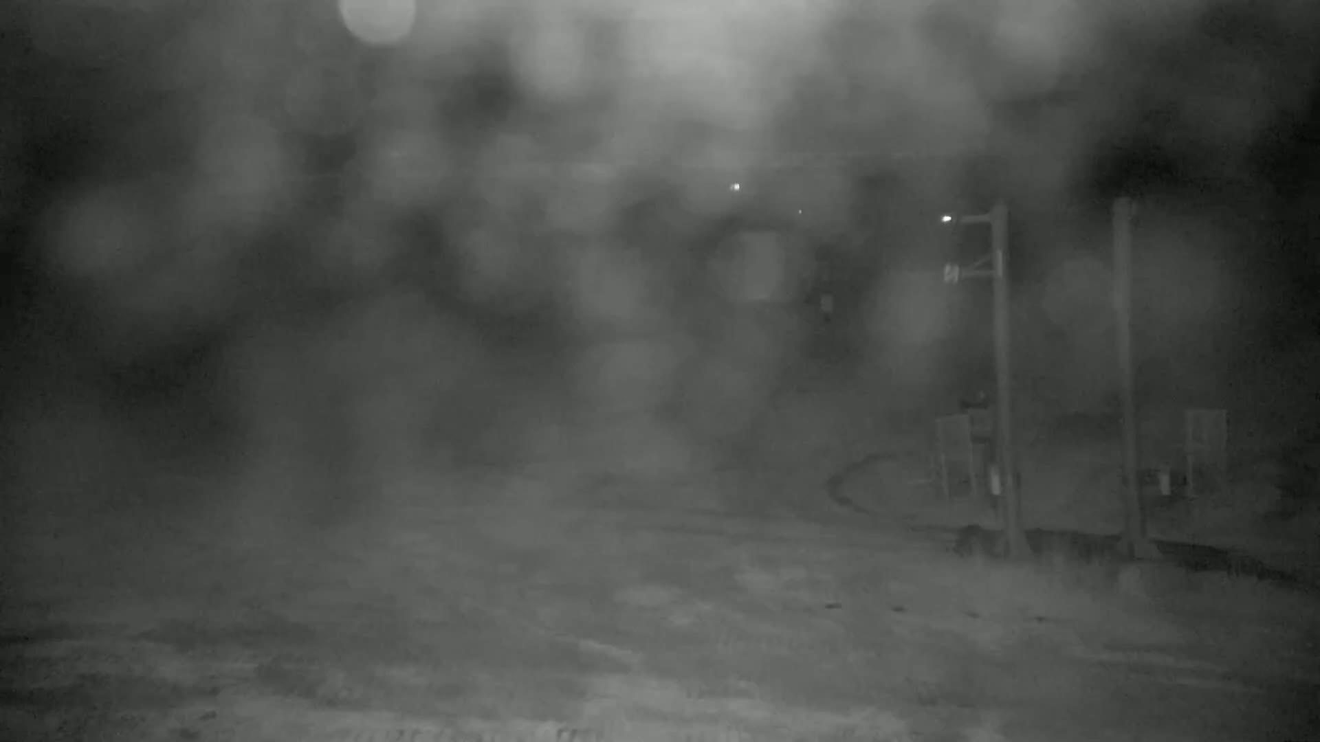 A blurry, nighttime outdoor scene shows a mostly empty, wet roadway with indistinct structures and light posts under heavy rain or fog.