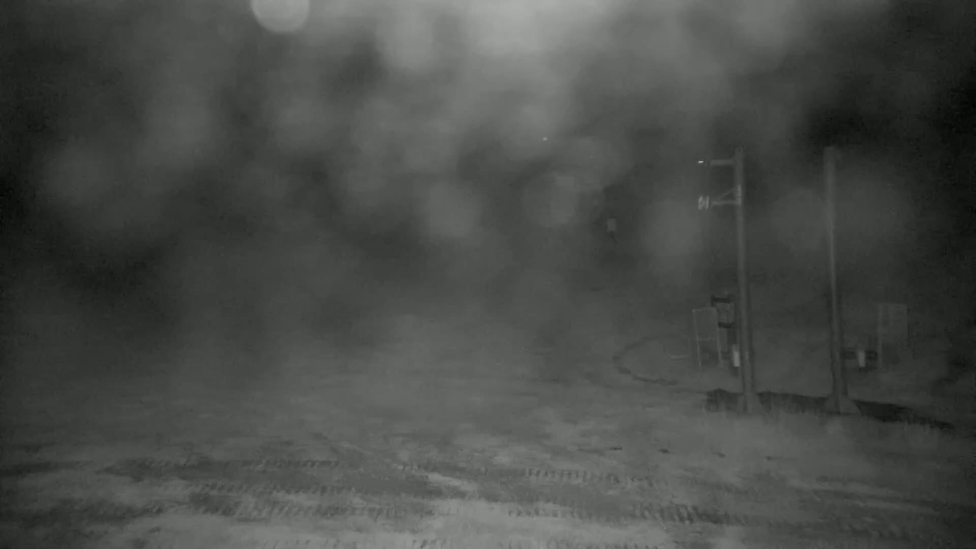 A blurry, nighttime image shows a snow-covered ground with indistinct objects and two tall poles in the heavy fog or snow.