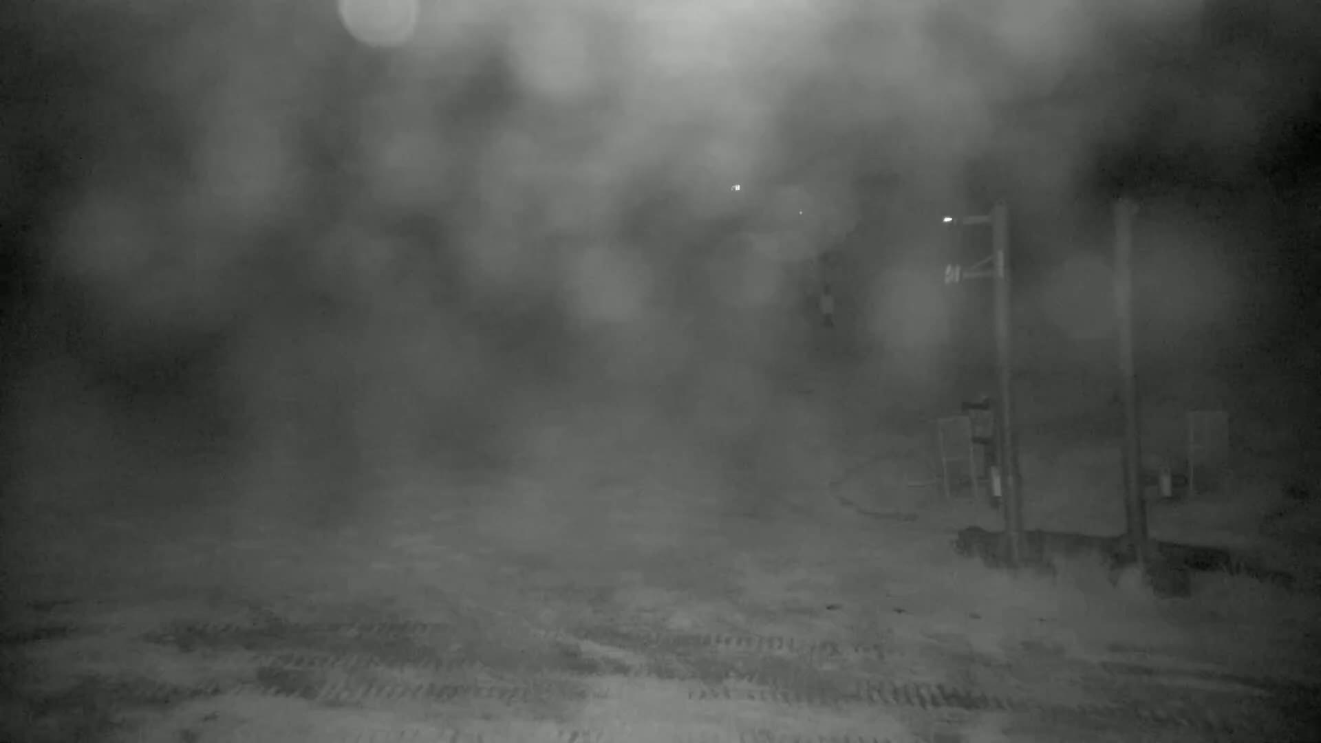 A blurry, nighttime scene shows a snow-covered area with indistinct structures and poles under heavy snow or fog.