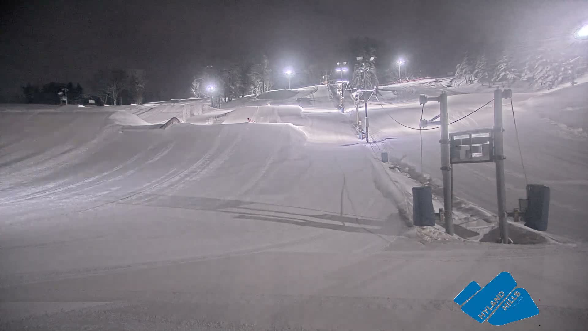 A brightly lit, snow-covered ski slope and terrain park with jumps and a tow rope are seen at night under a dark, cold sky with visible mist or light snow creating a hazy effect around the lights.
