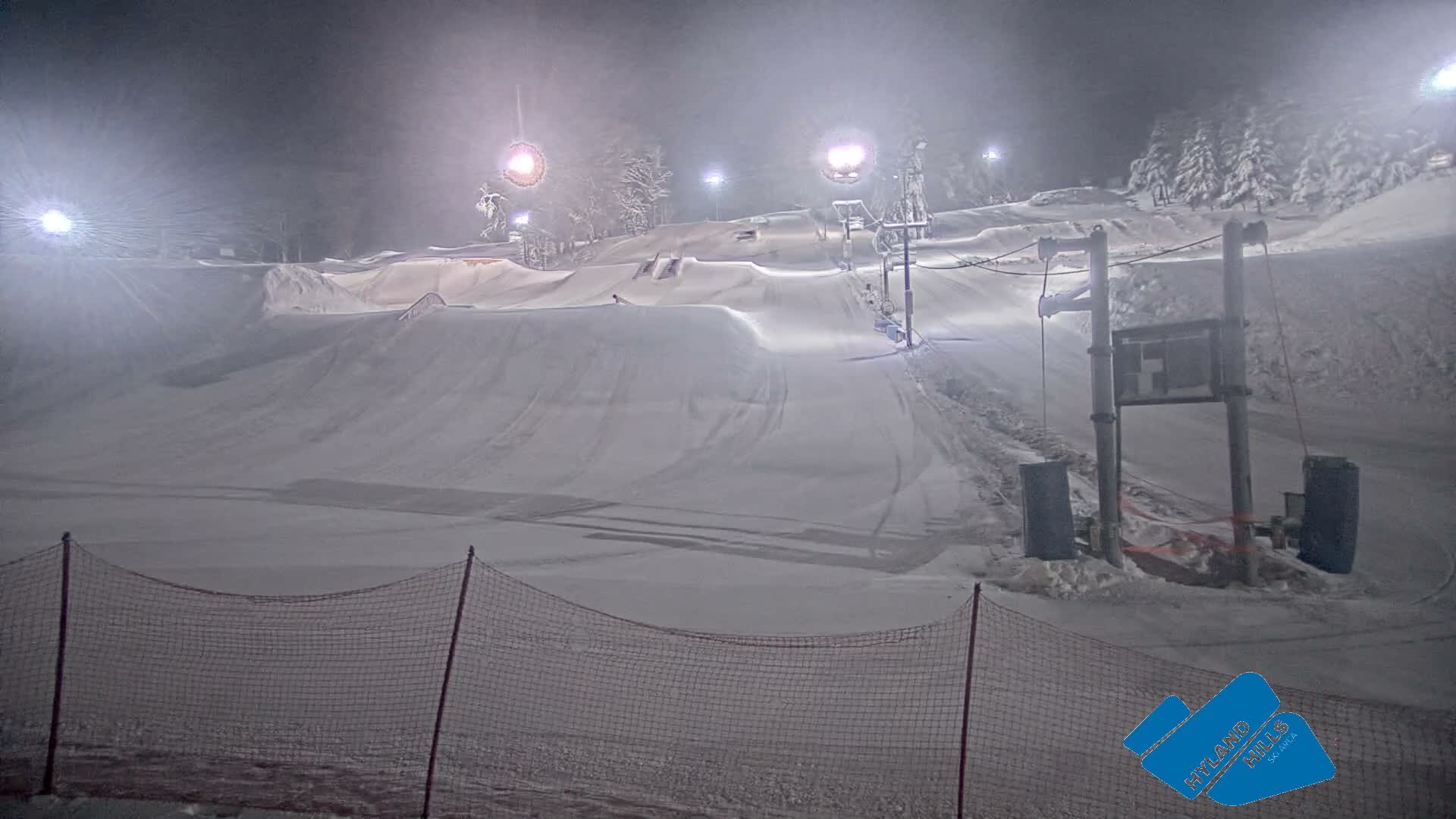 A brightly lit, snow-covered ski slope and terrain park with jumps and a tow rope are seen at night under a dark, cold sky with visible mist or light snow creating a hazy effect around the lights.