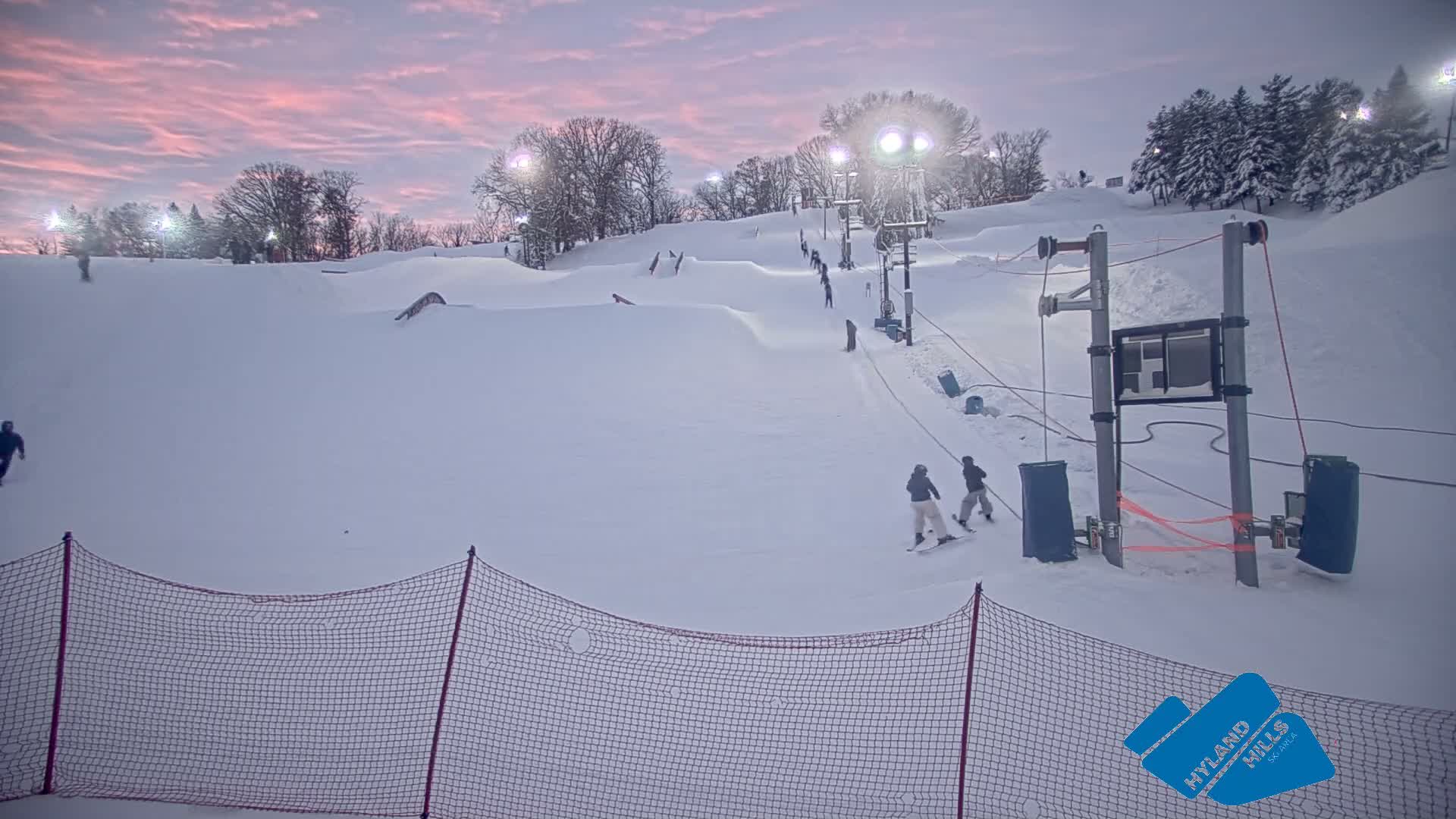 A brightly lit, snow-covered ski slope and terrain park with jumps and a tow rope are seen at night under a dark, cold sky with visible mist or light snow creating a hazy effect around the lights.