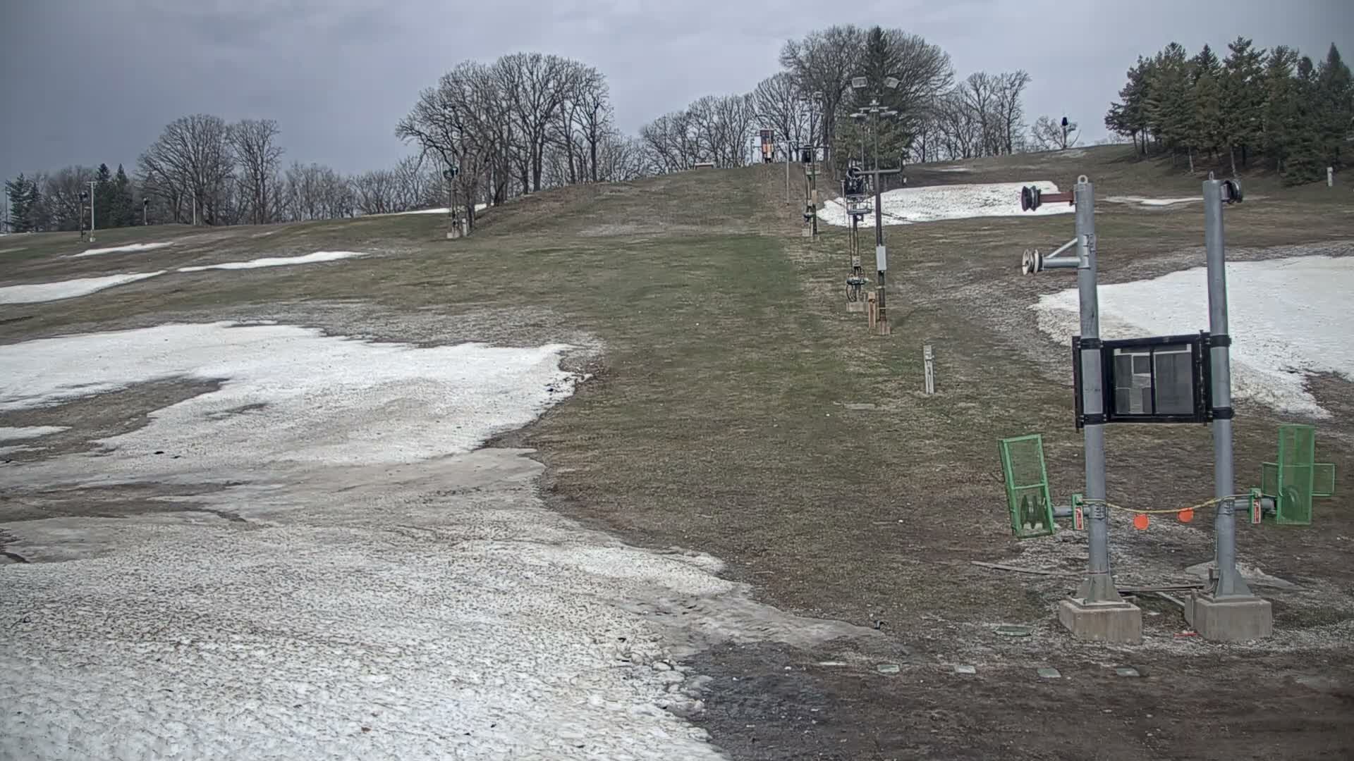 A brightly lit, snow-covered ski slope and terrain park with jumps and a tow rope are seen at night under a dark, cold sky with visible mist or light snow creating a hazy effect around the lights.