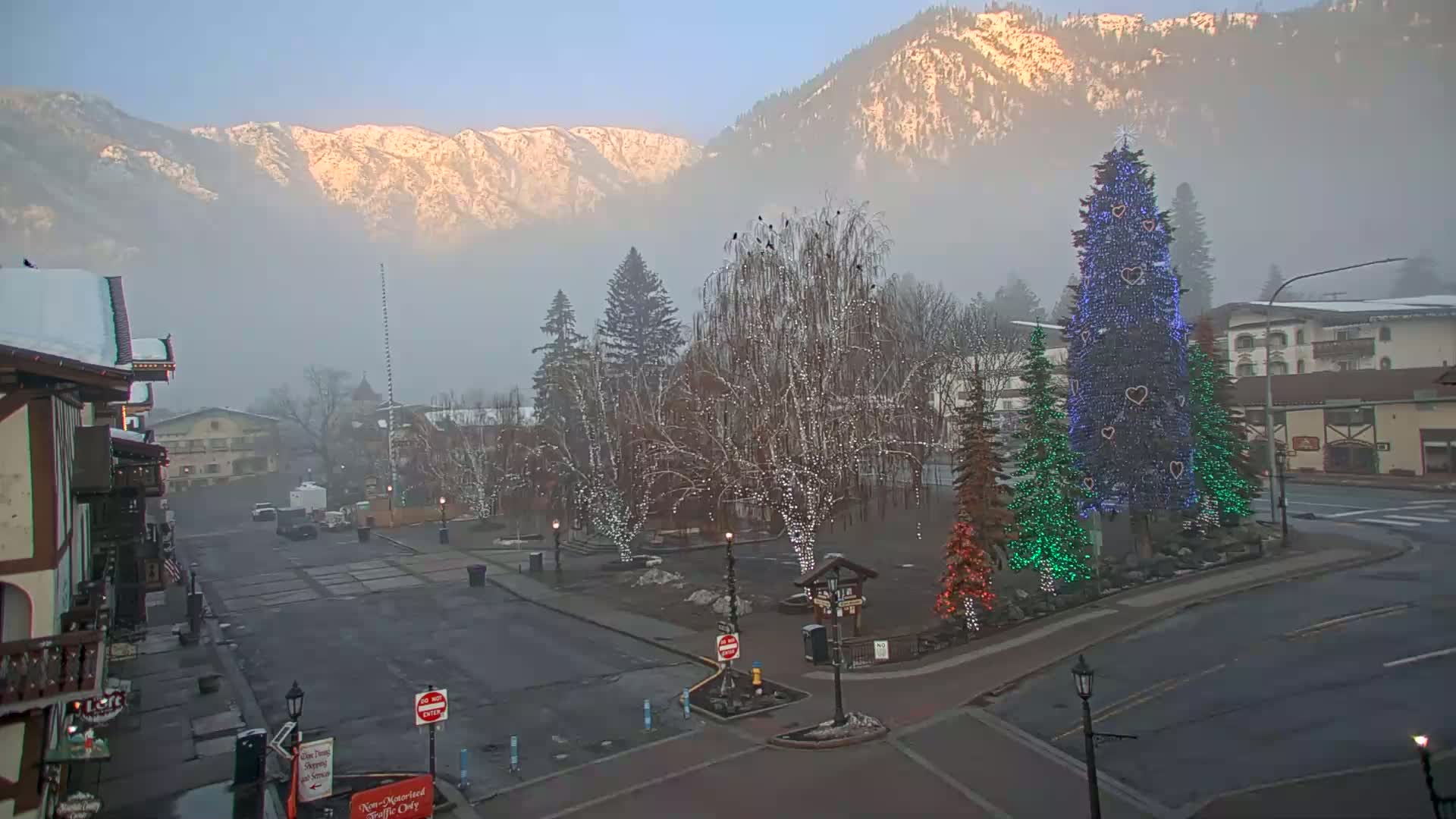 A foggy morning scene shows a town square with a large decorated Christmas tree and smaller trees with lights, in front of snow-covered mountains.
