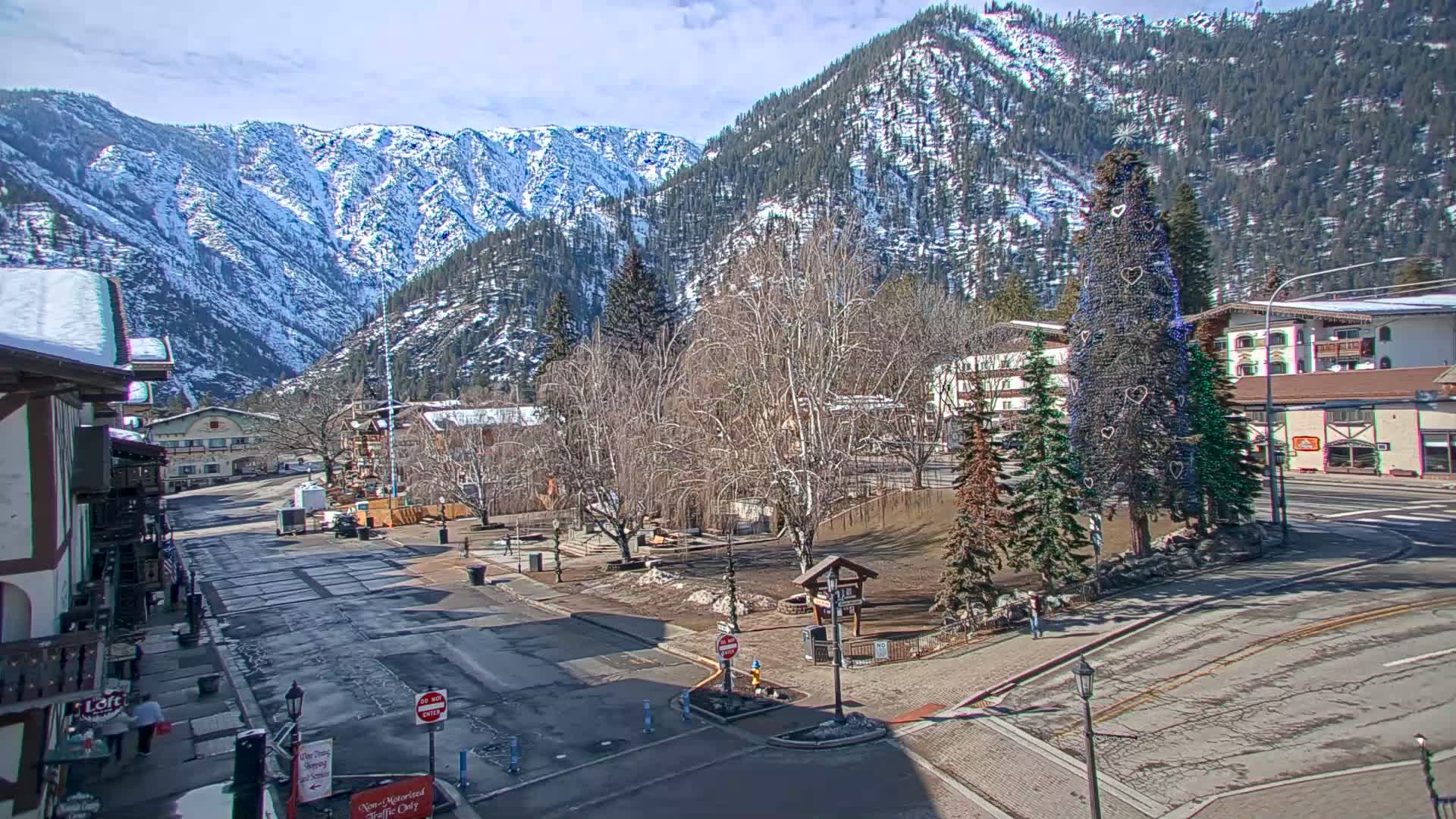 A mostly empty street in a mountain town with snow-capped mountains in the background on a partly sunny day, featuring a decorated Christmas tree in a small park.