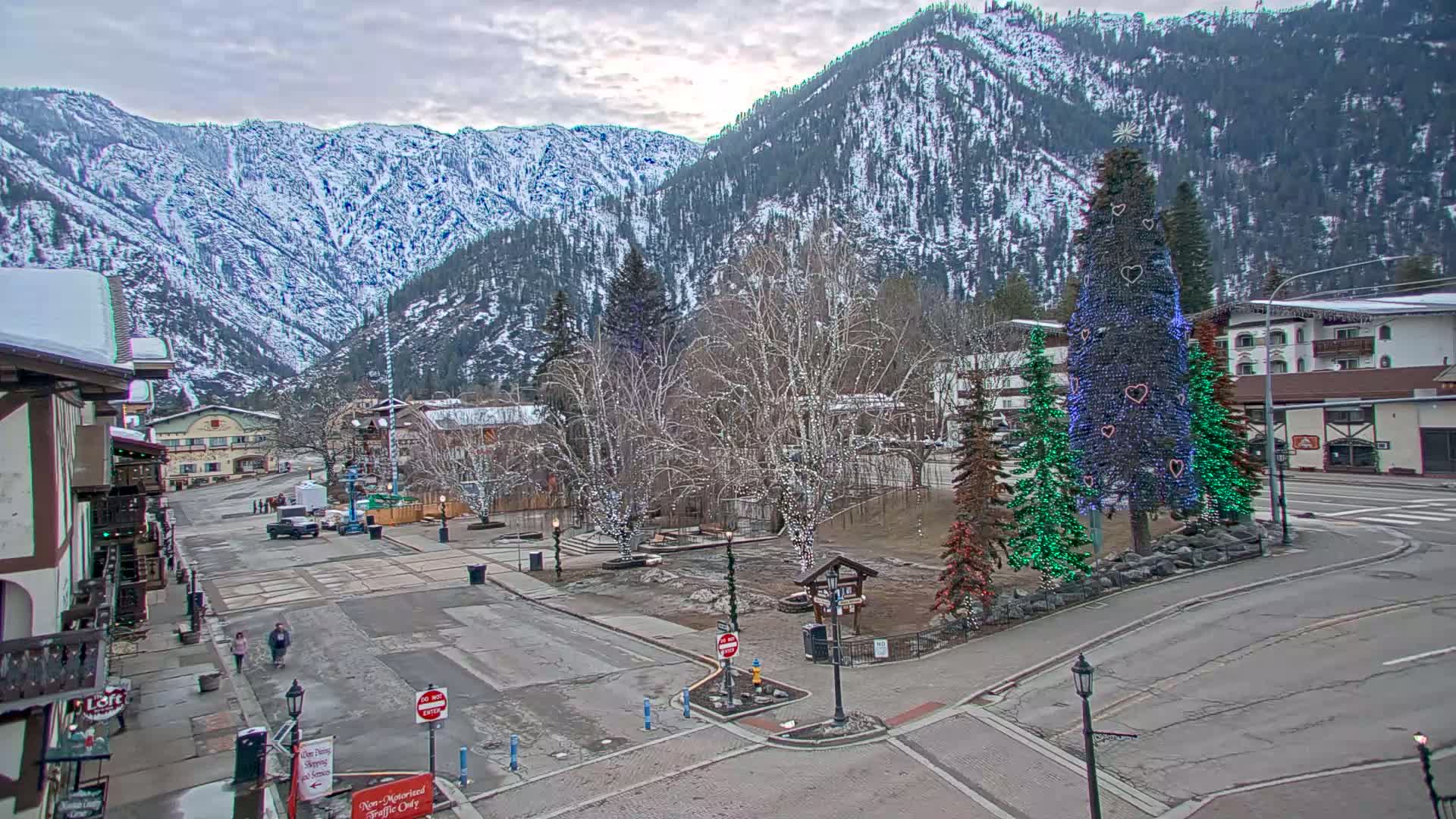 A snow-covered mountain range overlooks a town square with decorated Christmas trees, bare trees with string lights, and a few cars parked on the street under an overcast sky.