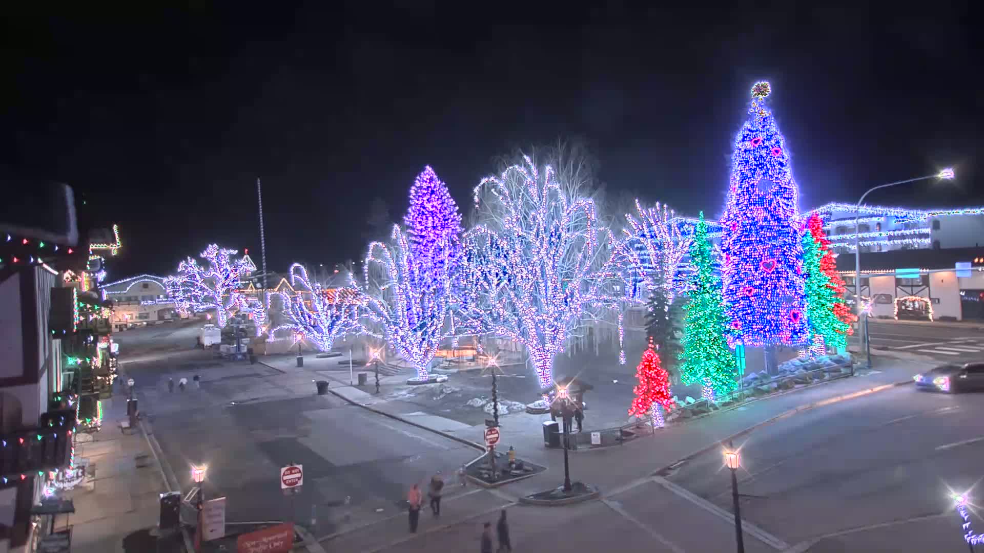 A town square at night is illuminated with colorful Christmas lights adorning trees and buildings.