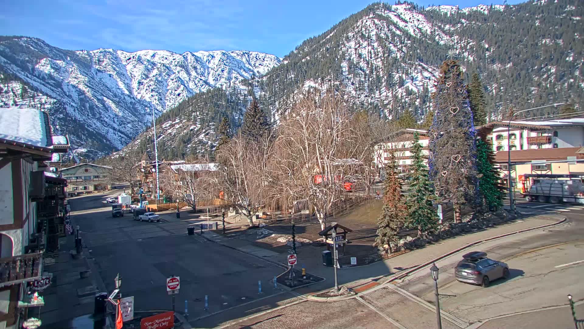 A snow-covered mountain range overlooks a town square with a large decorated Christmas tree, bare winter trees, and several cars under a sunny sky.