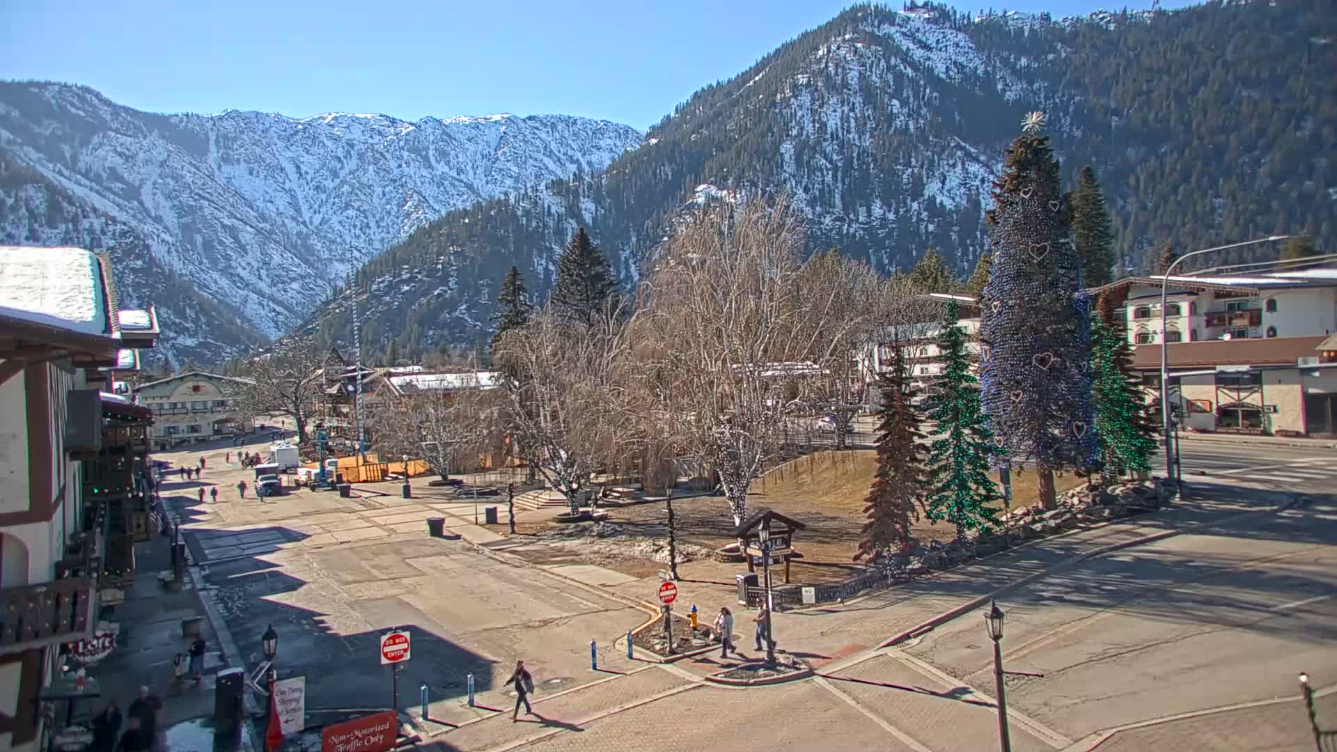A sunny day reveals a town square with decorated evergreen trees, snow-dusted mountains in the background, and a few pedestrians walking about.