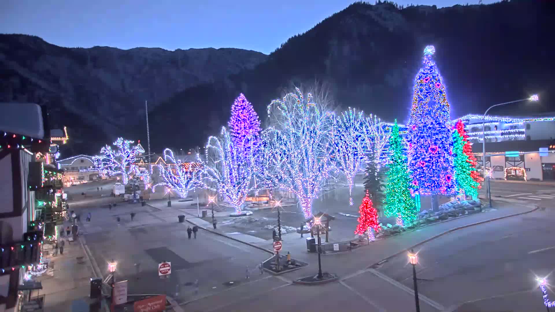 A town square at night is illuminated by brightly colored Christmas lights adorning trees and buildings, with snow-covered mountains in the background under a dark sky.