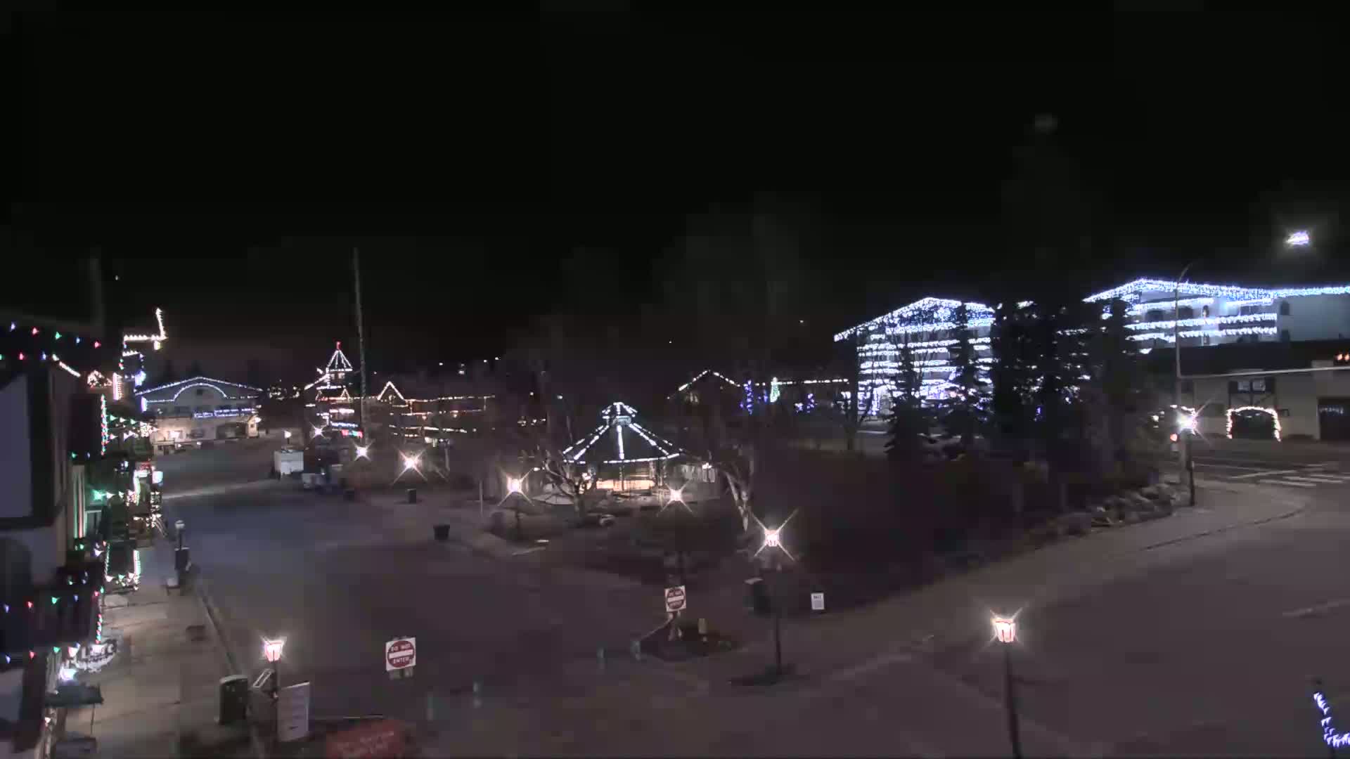 A nighttime view of a town square adorned with Christmas lights, featuring a gazebo and buildings decorated for the holidays under a clear, dark sky.