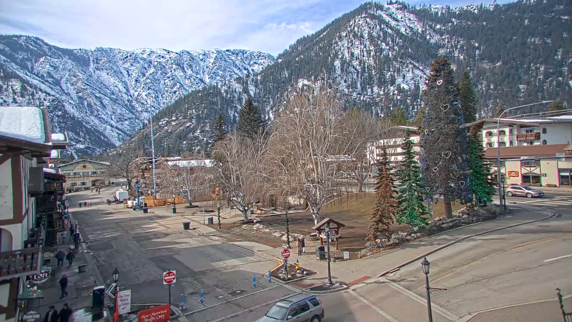 A snow-dusted mountain range overlooks a town square with a decorated evergreen tree, under a partly sunny sky.