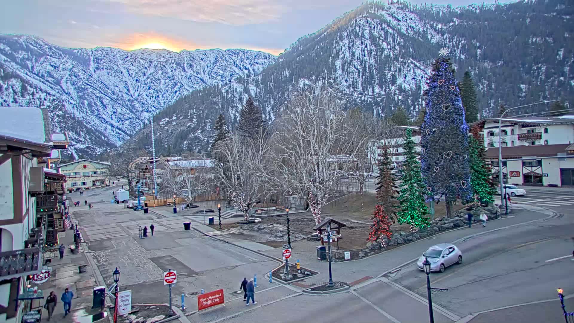 A snow-covered mountain town at sunset, featuring a large, illuminated Christmas tree in a town square, with pedestrians and a few cars visible.