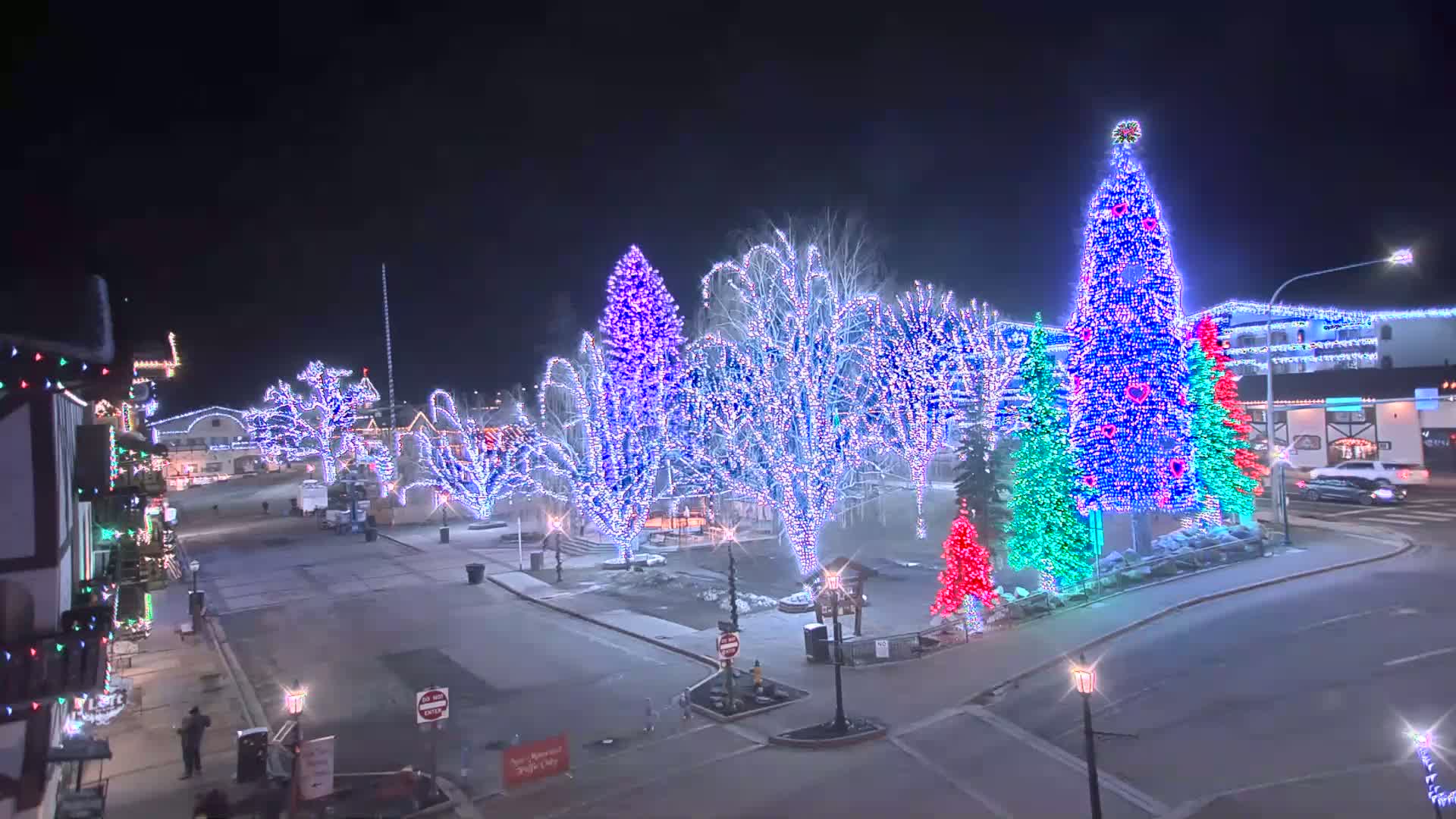 A town square at night is decorated with colorful Christmas lights strung on trees and shaped into large, illuminated Christmas trees.