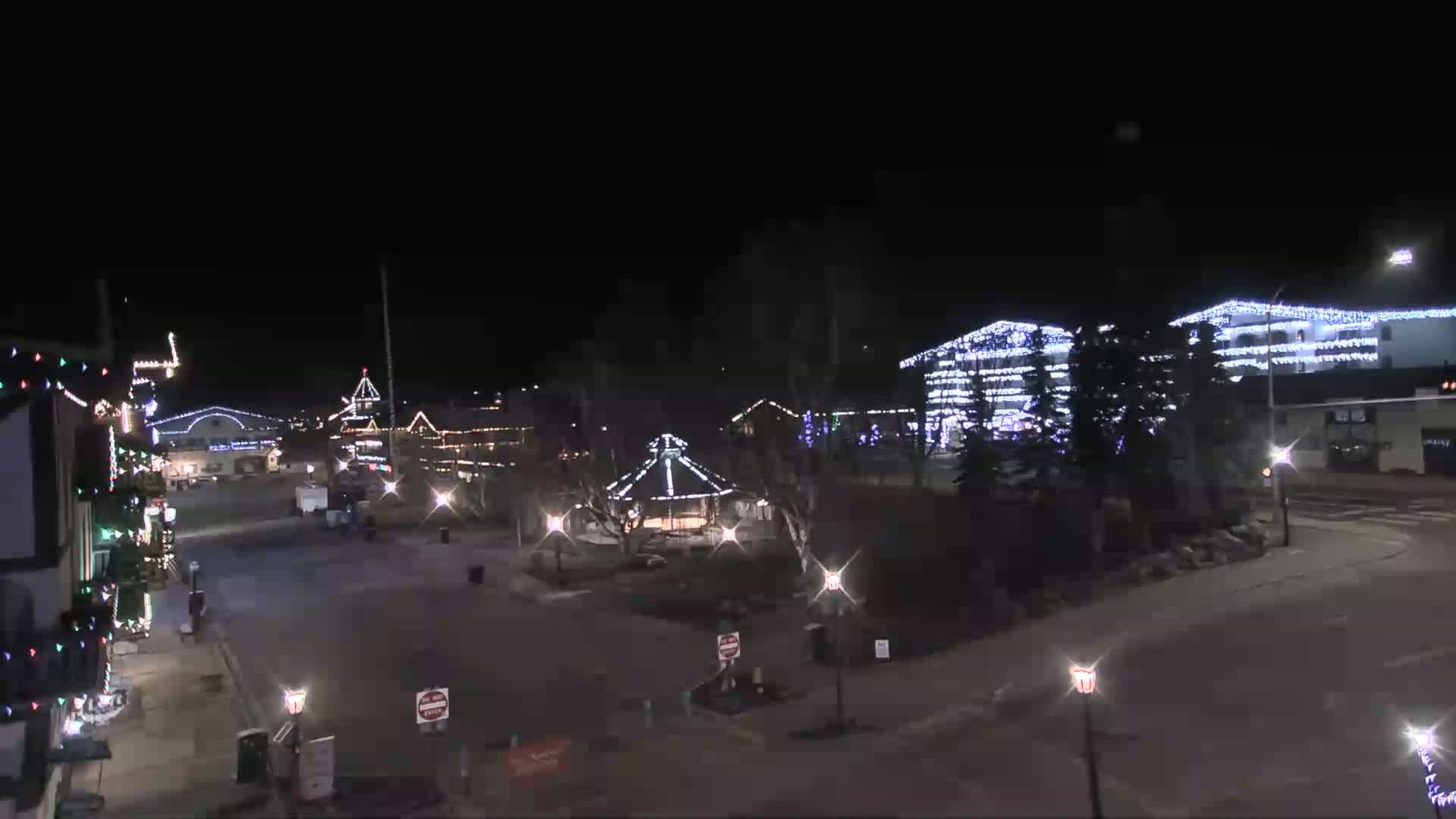 A nighttime view of a town square decorated with Christmas lights, featuring buildings, a gazebo, and streetlights under a clear, dark sky.