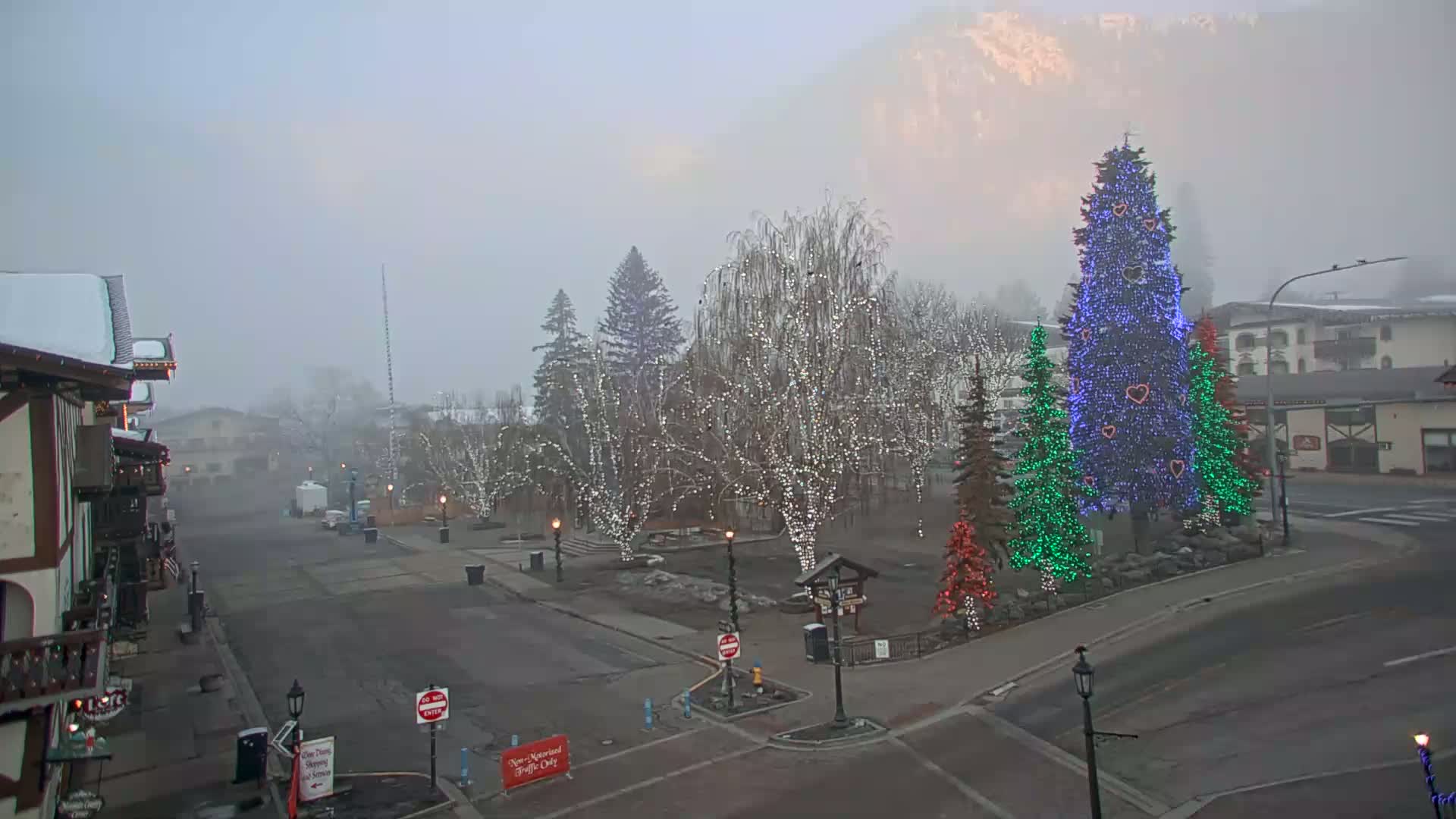 A foggy town square features several trees decorated with Christmas lights, including a large blue tree in the center, with snow on some buildings and the ground.