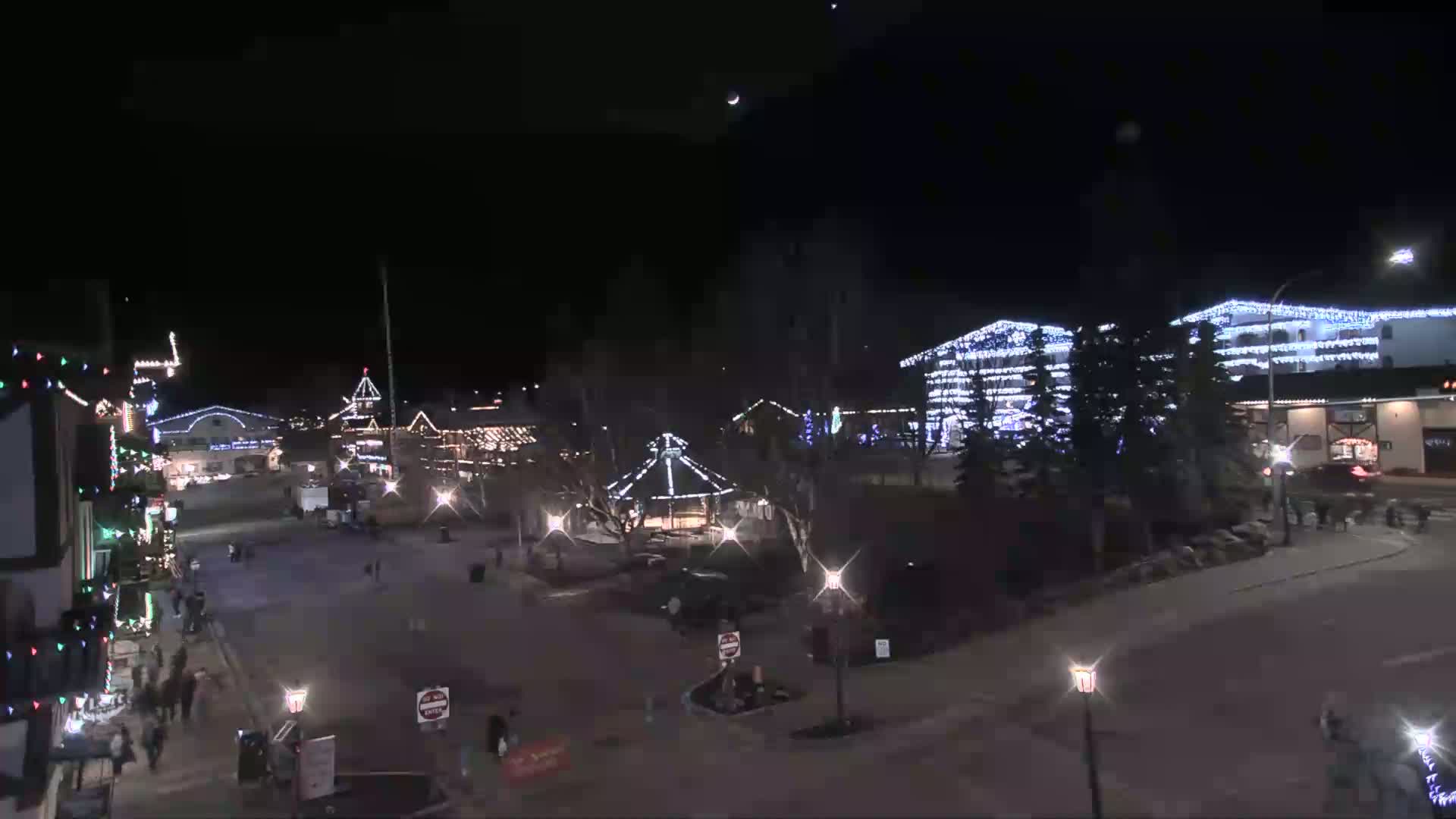 A nighttime, snow-covered town square is illuminated by strings of lights on buildings and lampposts, with a gazebo at its center.