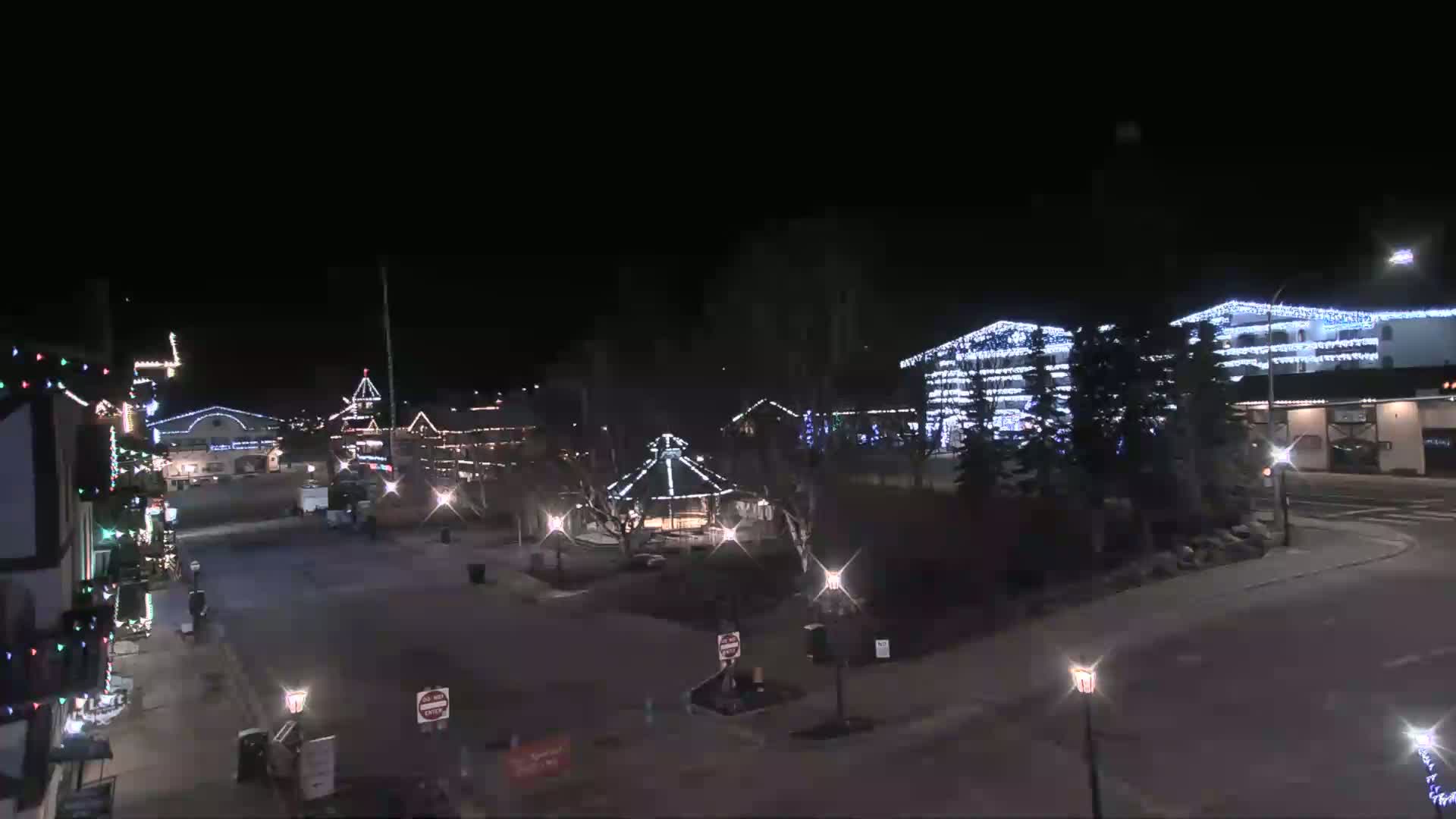 A nighttime view of a town square adorned with Christmas lights, featuring buildings, a gazebo, and sparsely populated streets.