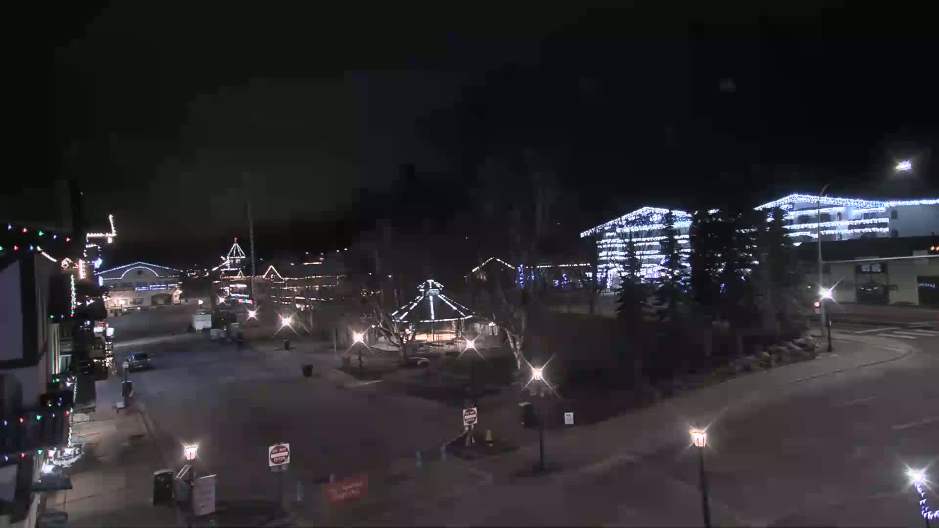 A nighttime view of a town square with buildings decorated with Christmas lights, a gazebo in the center, and a mostly empty street.