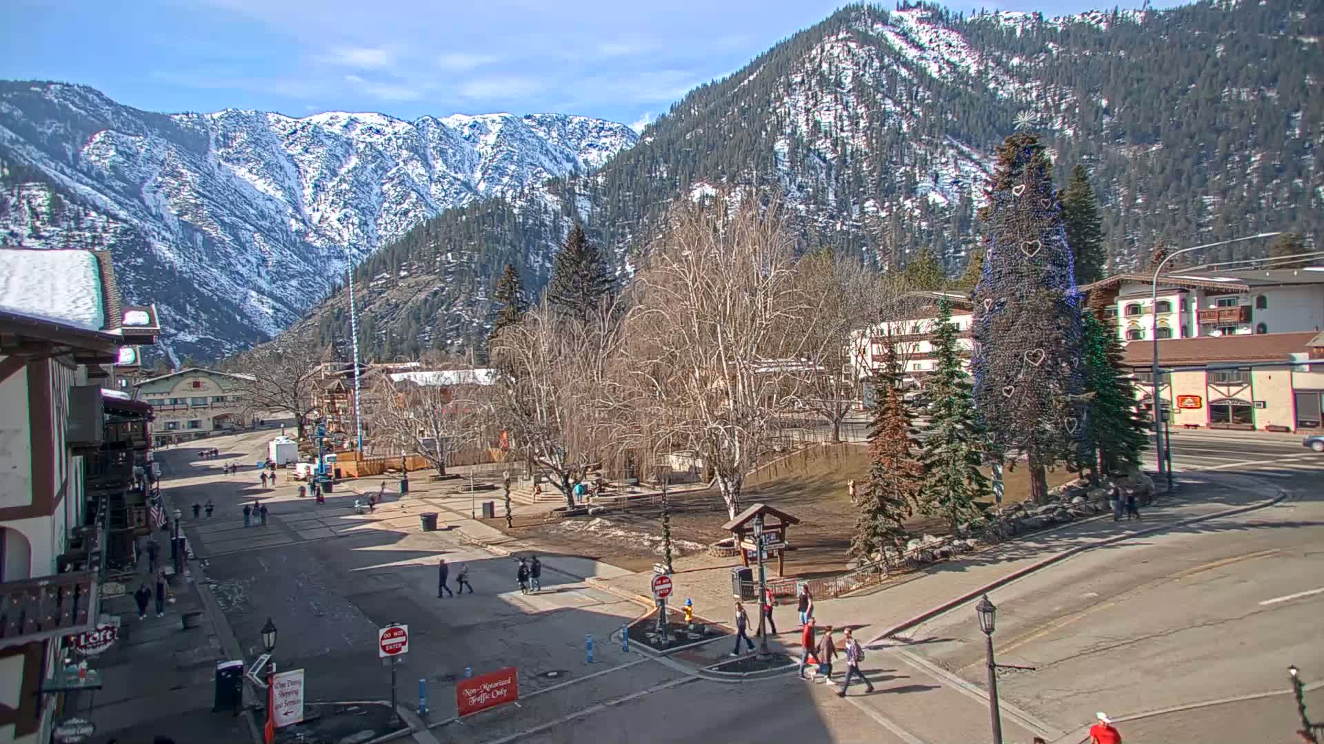 A town square with a decorated Christmas tree is situated in front of snow-capped mountains under a clear, sunny sky.