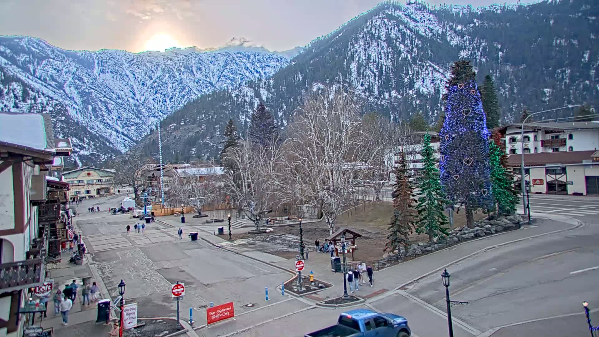 A town square with a decorated evergreen tree in the center is nestled at the base of snow-covered mountains under a partly sunny sky.