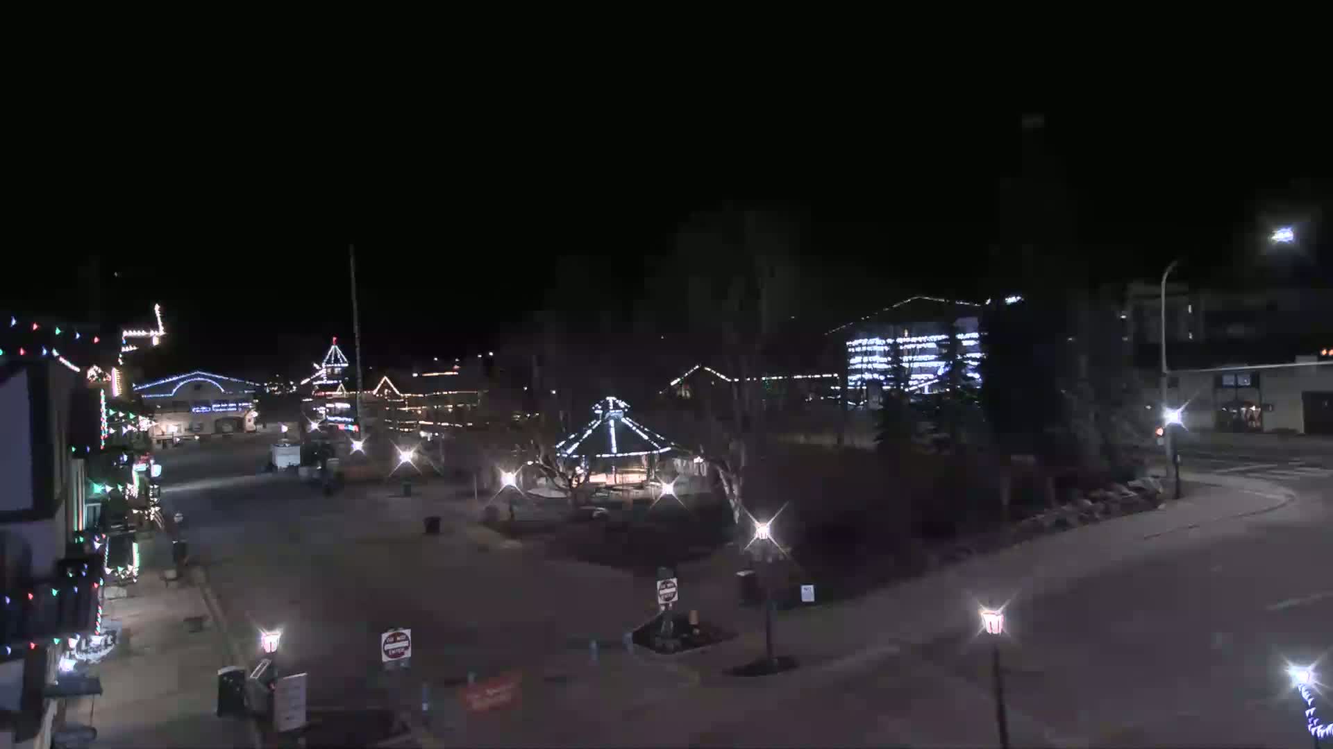 A nighttime view of a town square adorned with Christmas lights, featuring a gazebo in the center and buildings decorated with lights along the edges.