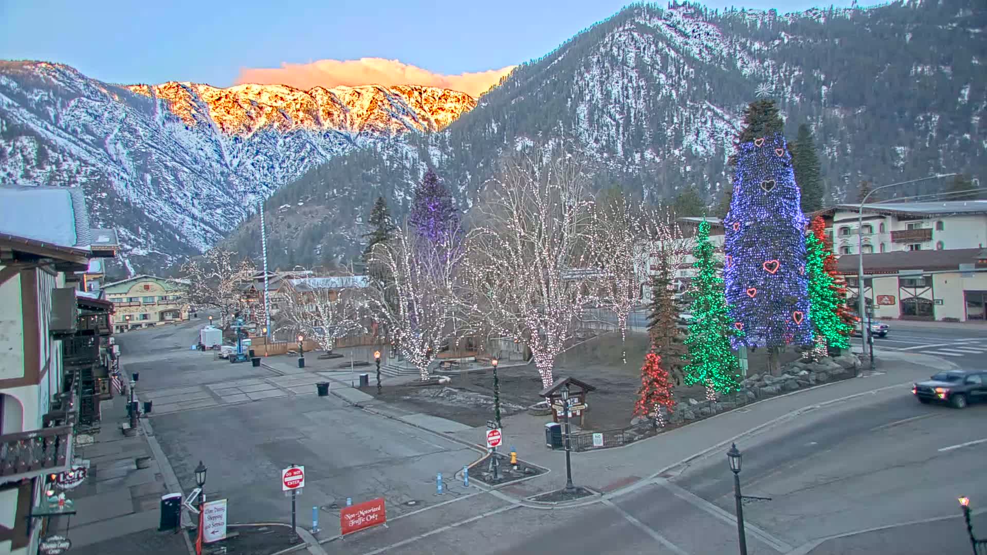 A snow-covered mountain town square at dusk, illuminated by strings of lights on trees and large decorated Christmas trees, with a few cars on the street.