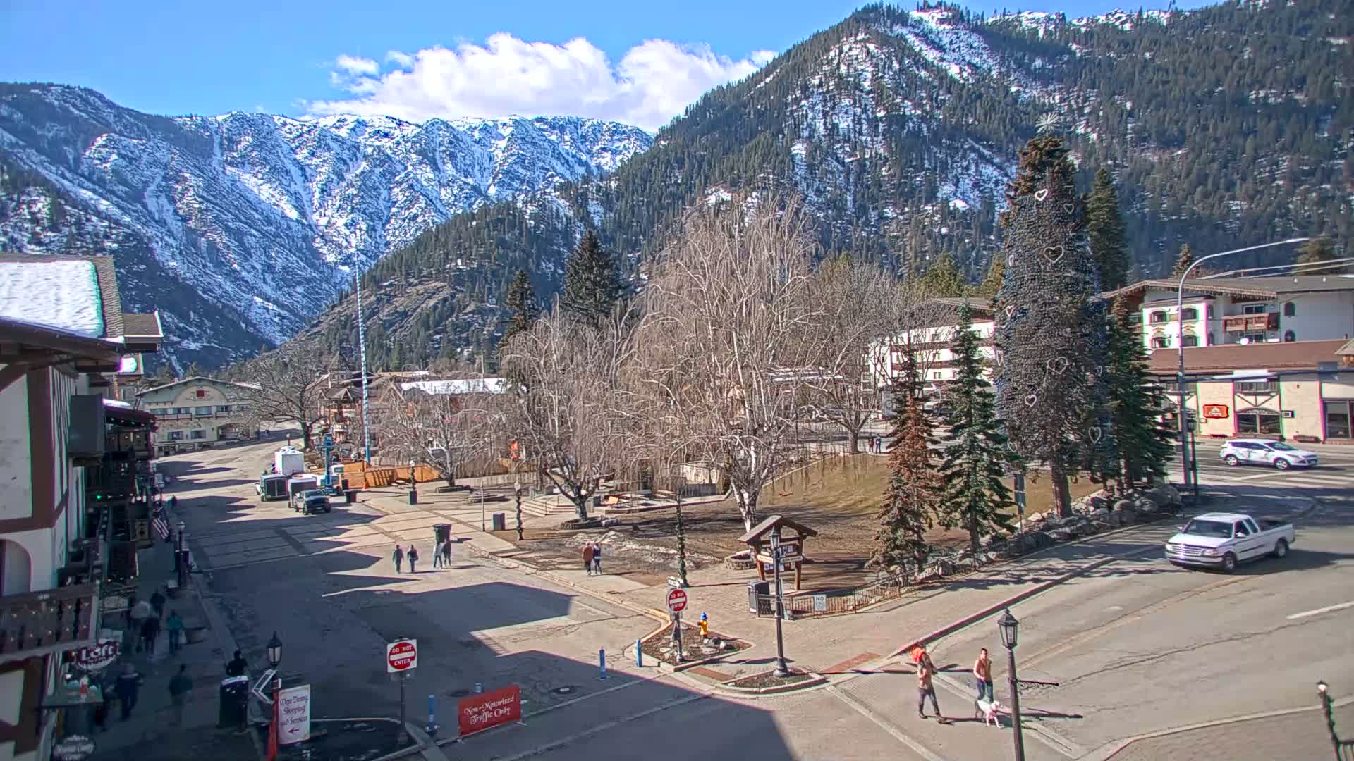 A mostly sunny day reveals a town square with a large decorated evergreen tree, surrounded by buildings and snow-capped mountains in the background.