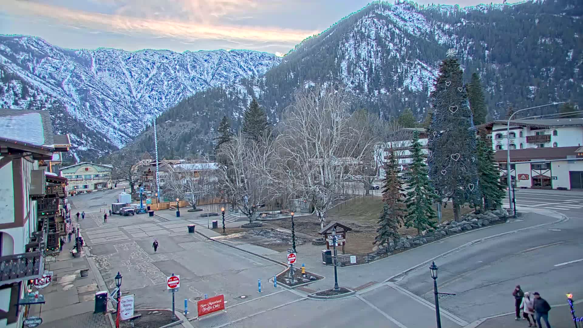 A snow-dusted mountain range overlooks a town square with a decorated evergreen tree, bare deciduous trees, and a few pedestrians, under a mostly cloudy sky.