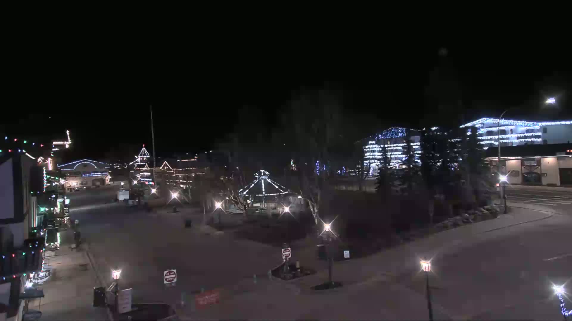 A nighttime view of a town square with buildings decorated with Christmas lights, a gazebo in the center, and a mostly empty street.
