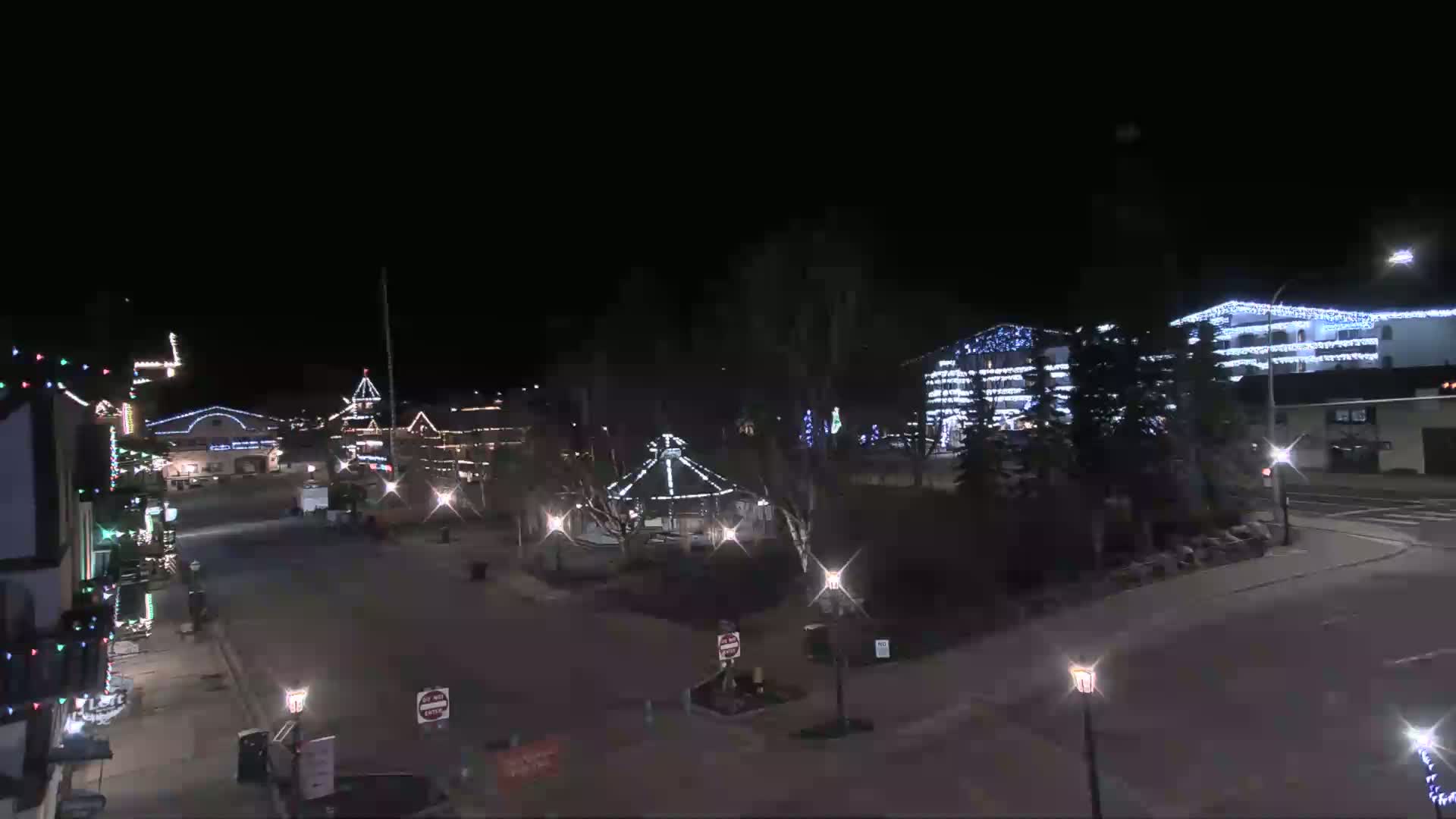 A nighttime view of a town square decorated with Christmas lights, featuring buildings, a gazebo, and streetlights under a clear, dark sky.