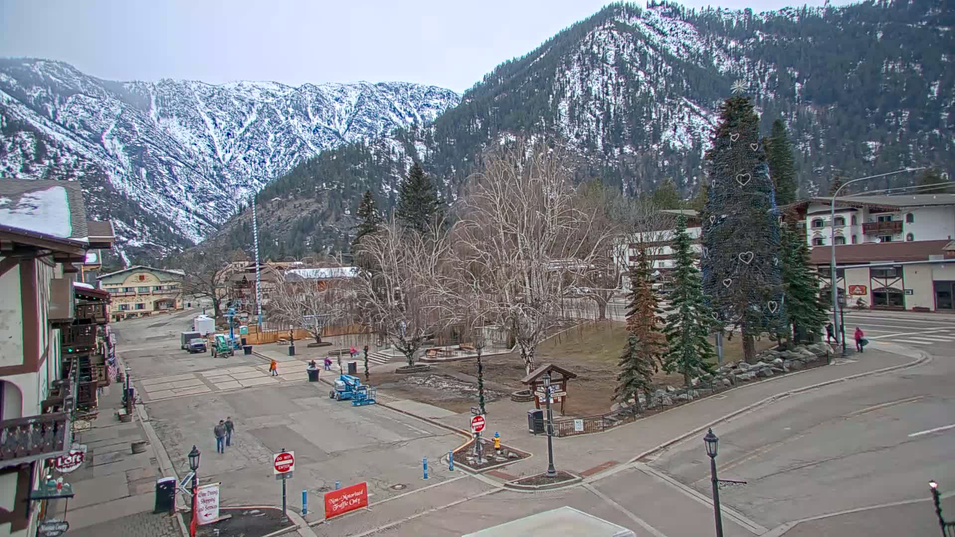 A mostly clear day shows a town square nestled in a valley between snow-capped mountains, with a decorated evergreen tree in the center.