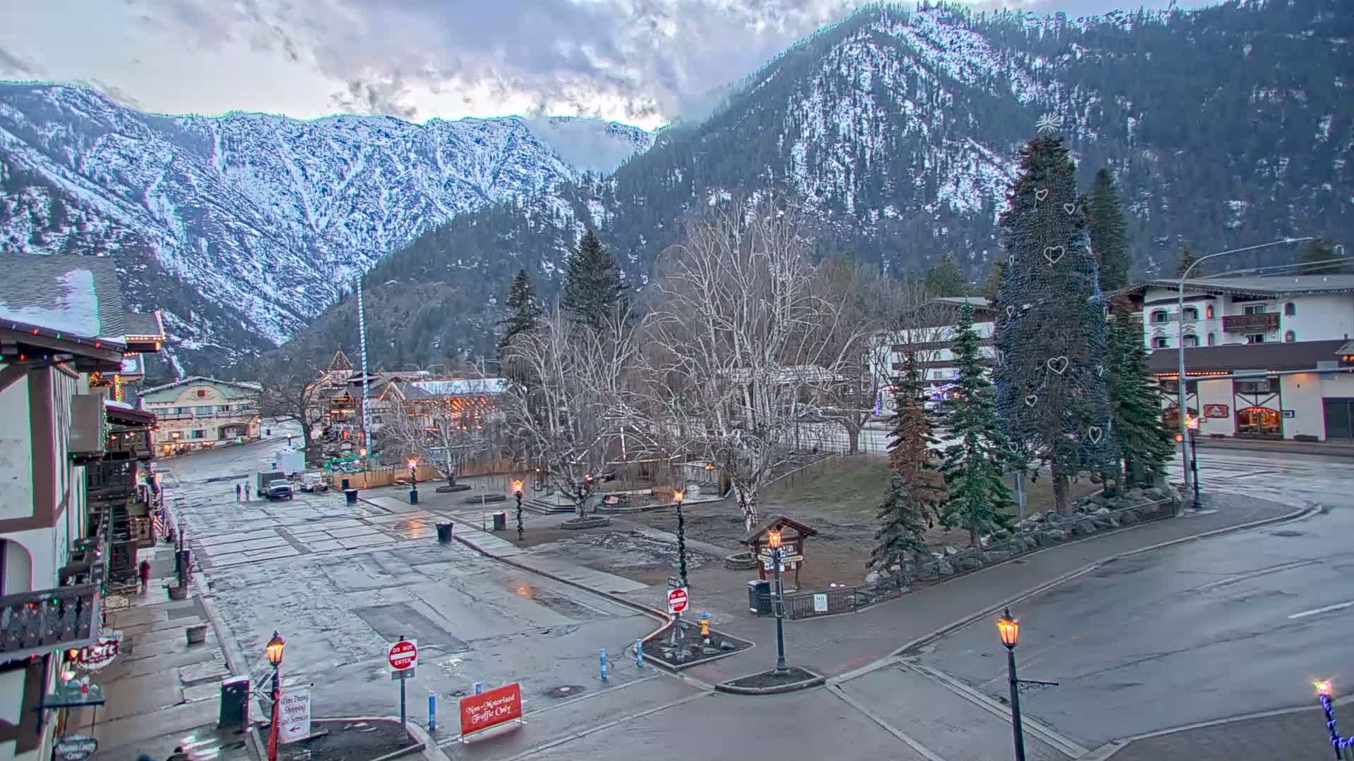 A wet town square sits below snow-covered mountains under an overcast sky, with a decorated Christmas tree in the foreground.