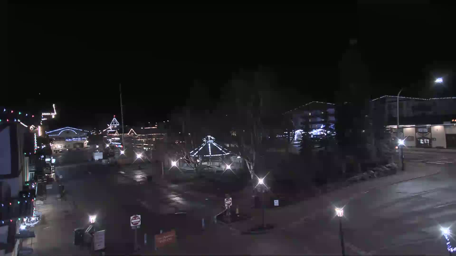 A nighttime view of a town square with buildings decorated with lights, a gazebo in the center, and a few cars parked along the street.