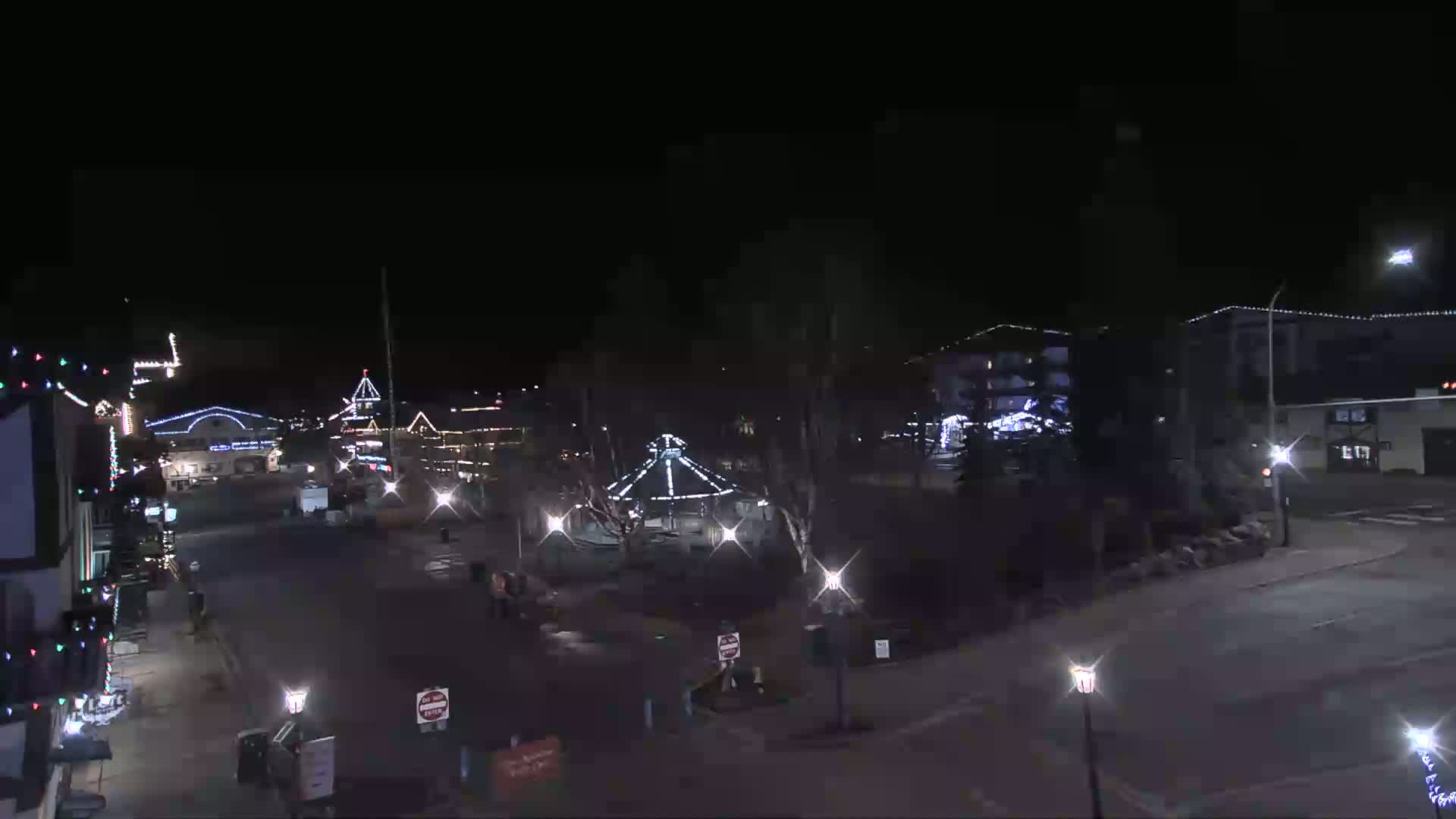 A nighttime view of a town square with buildings decorated in holiday lights, a gazebo in the center, and a mostly empty street.
